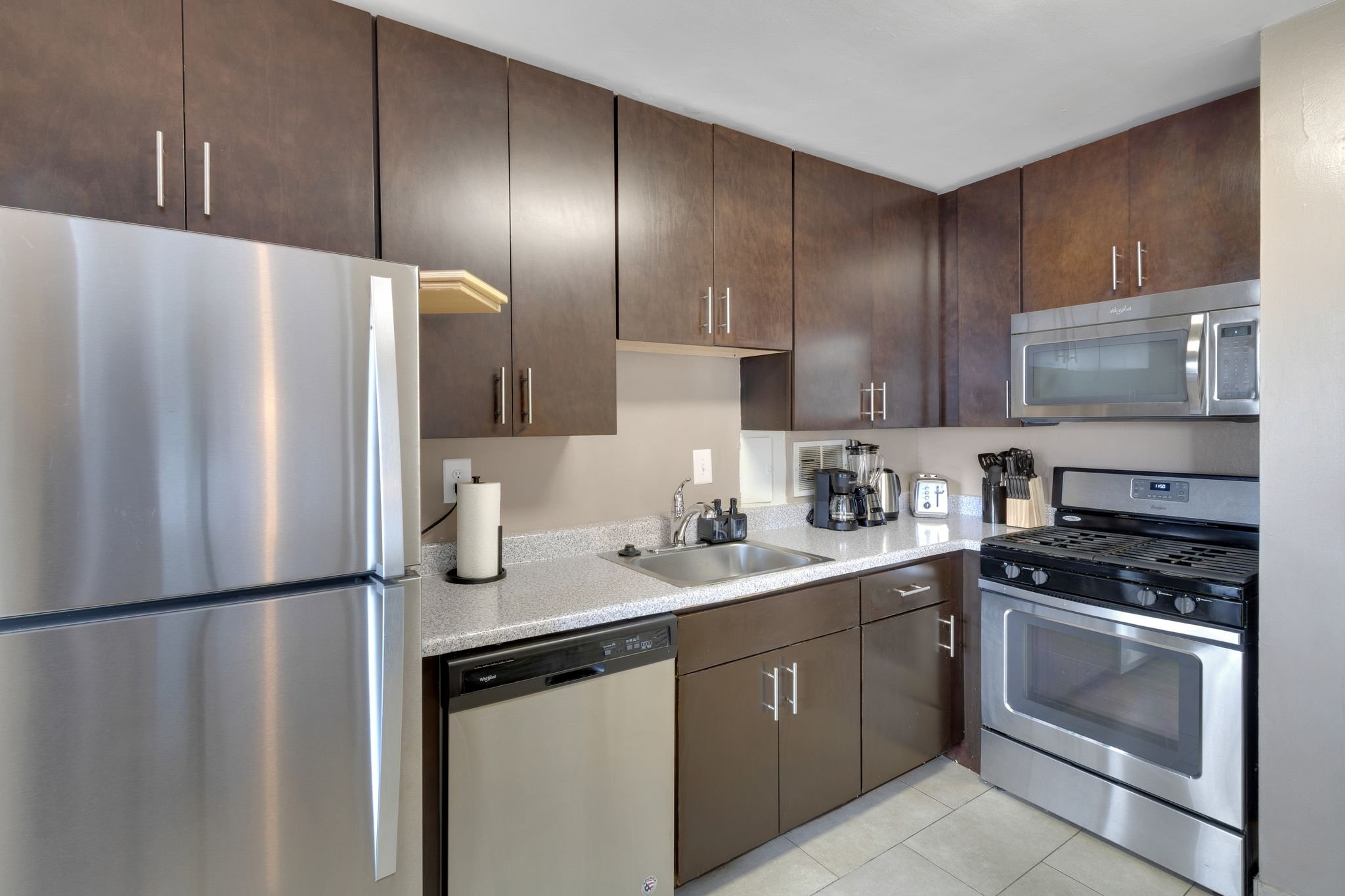 Modern kitchen with dark brown upper and lower cabinets, stainless steel refrigerator, microwave, stove, and small appliances on the countertop.