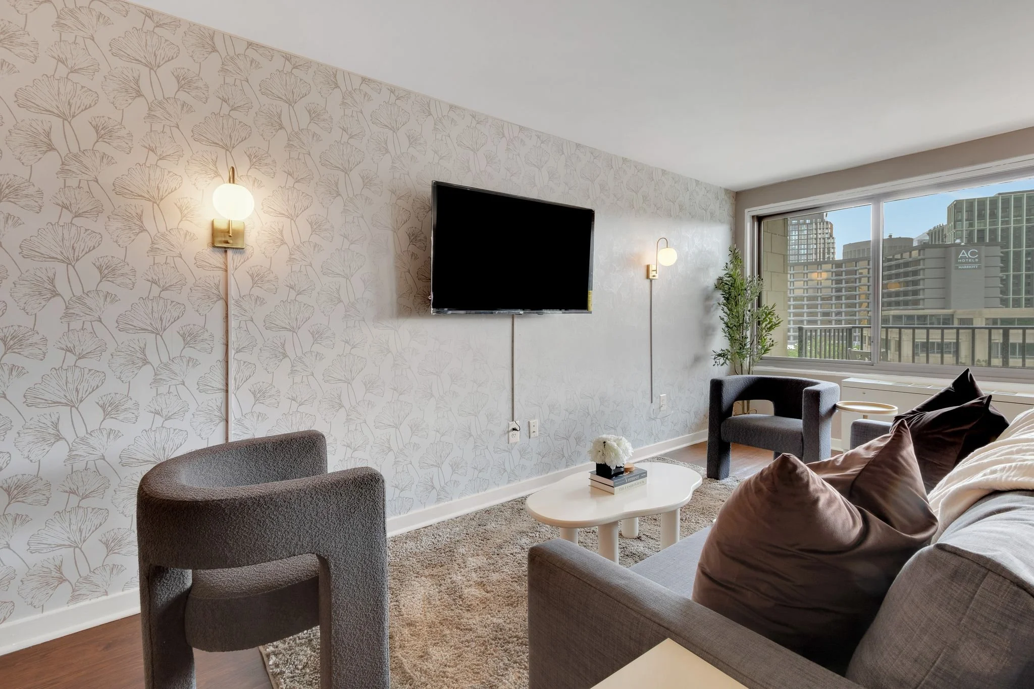 Living room with gray sofa, brown and black pillows, white coffee table with books and flowers, gray armchairs, large window with city view, patterned wallpaper, wall sconces, potted plant.