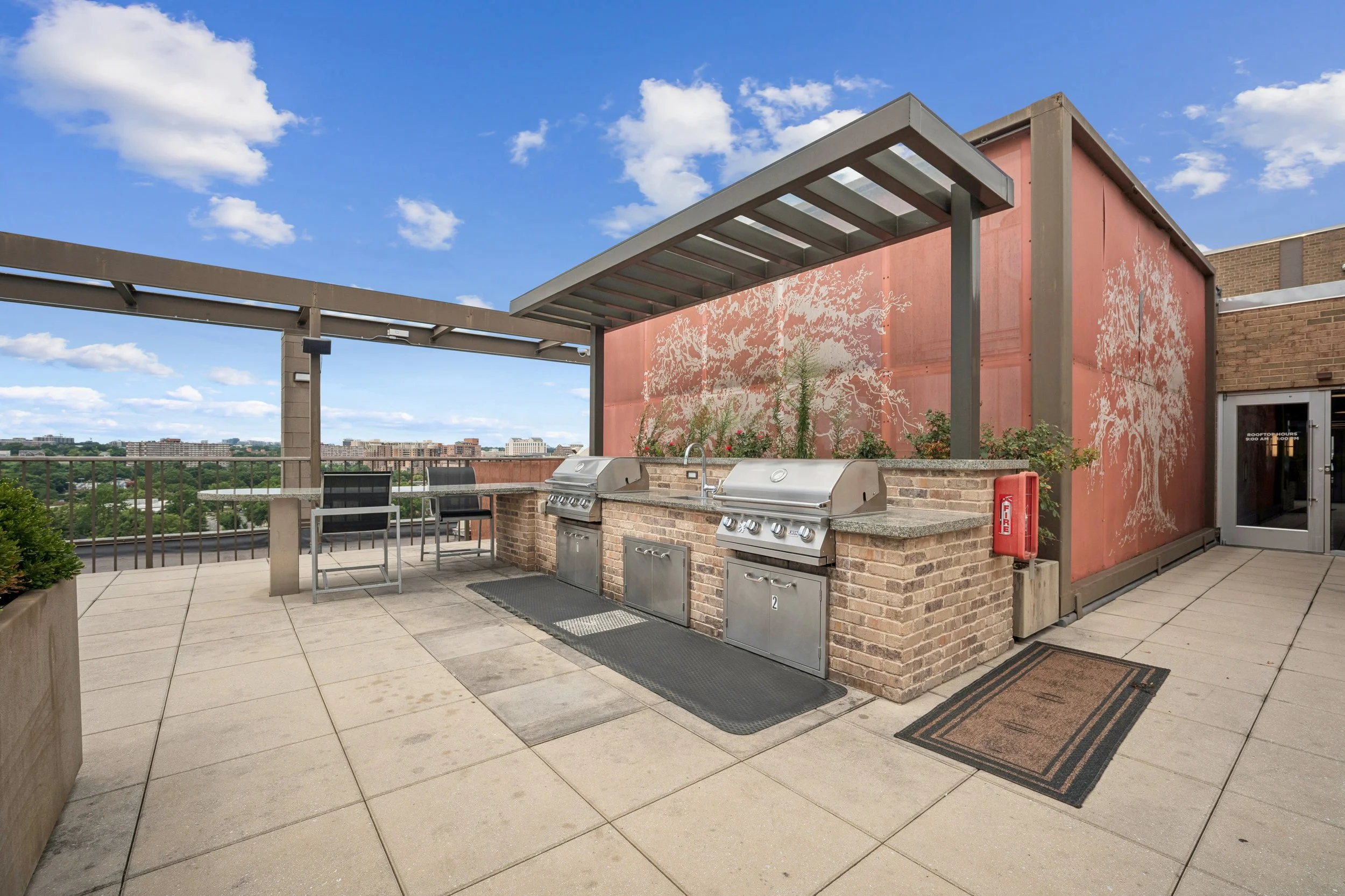 Rooftop outdoor grill area with two stainless steel grills, a sink, seating, and city skyline view under a blue sky with clouds.