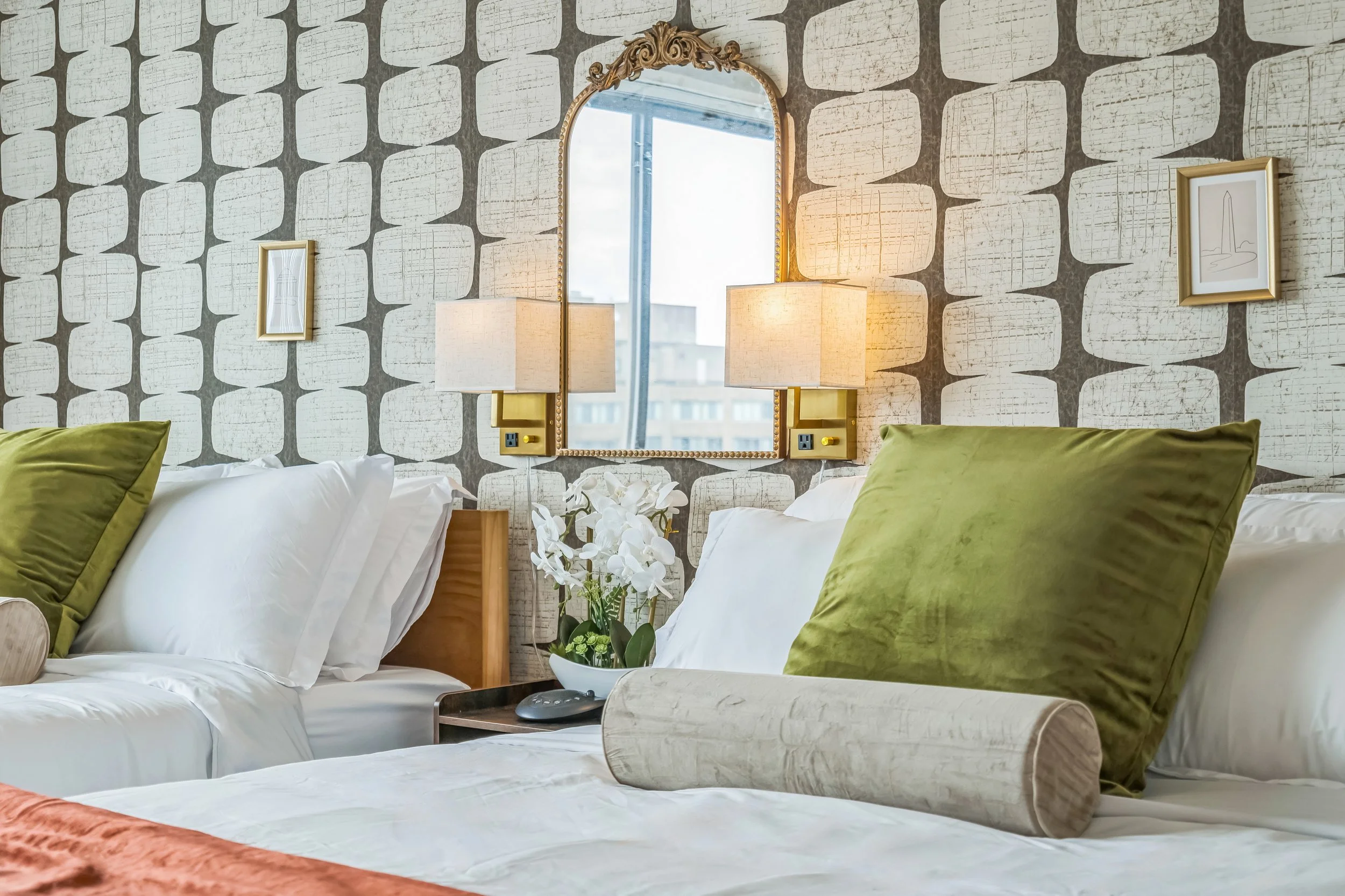 A cozy hotel bedroom with two beds, each with white linens, green pillows and beige bolster cushions. A vanity mirror with gold trim hangs on a patterned wallpapered wall, flanked by two wall lamps. There is a small black bedside table with a remote, and a white flower arrangement between the beds.