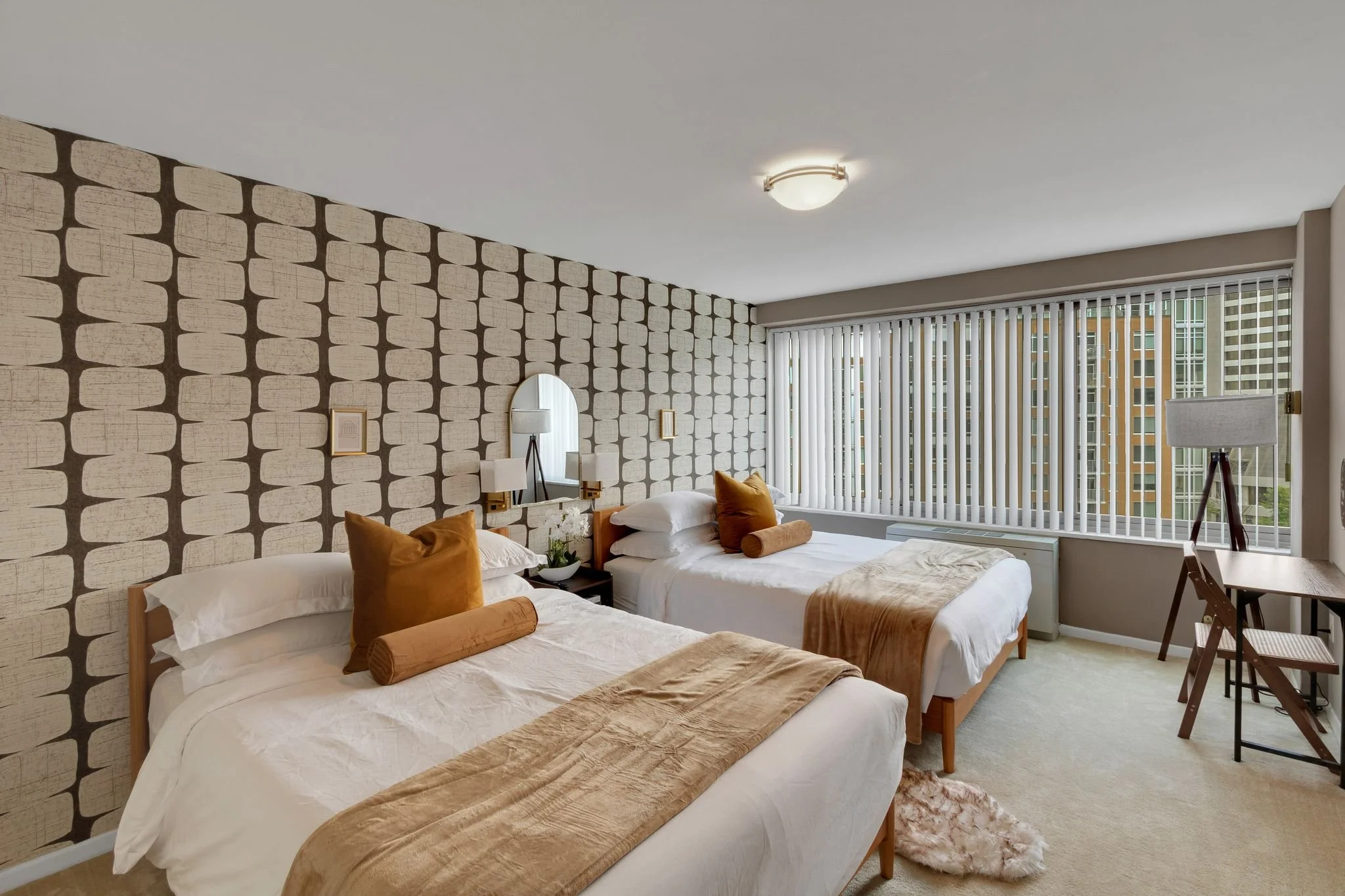 A bedroom with two beds, each with white bedding and brown pillows, a patterned accent wall, a large window with vertical blinds, a small black nightstand with a plant and a mirror, and a workspace with a wooden desk and a chair.