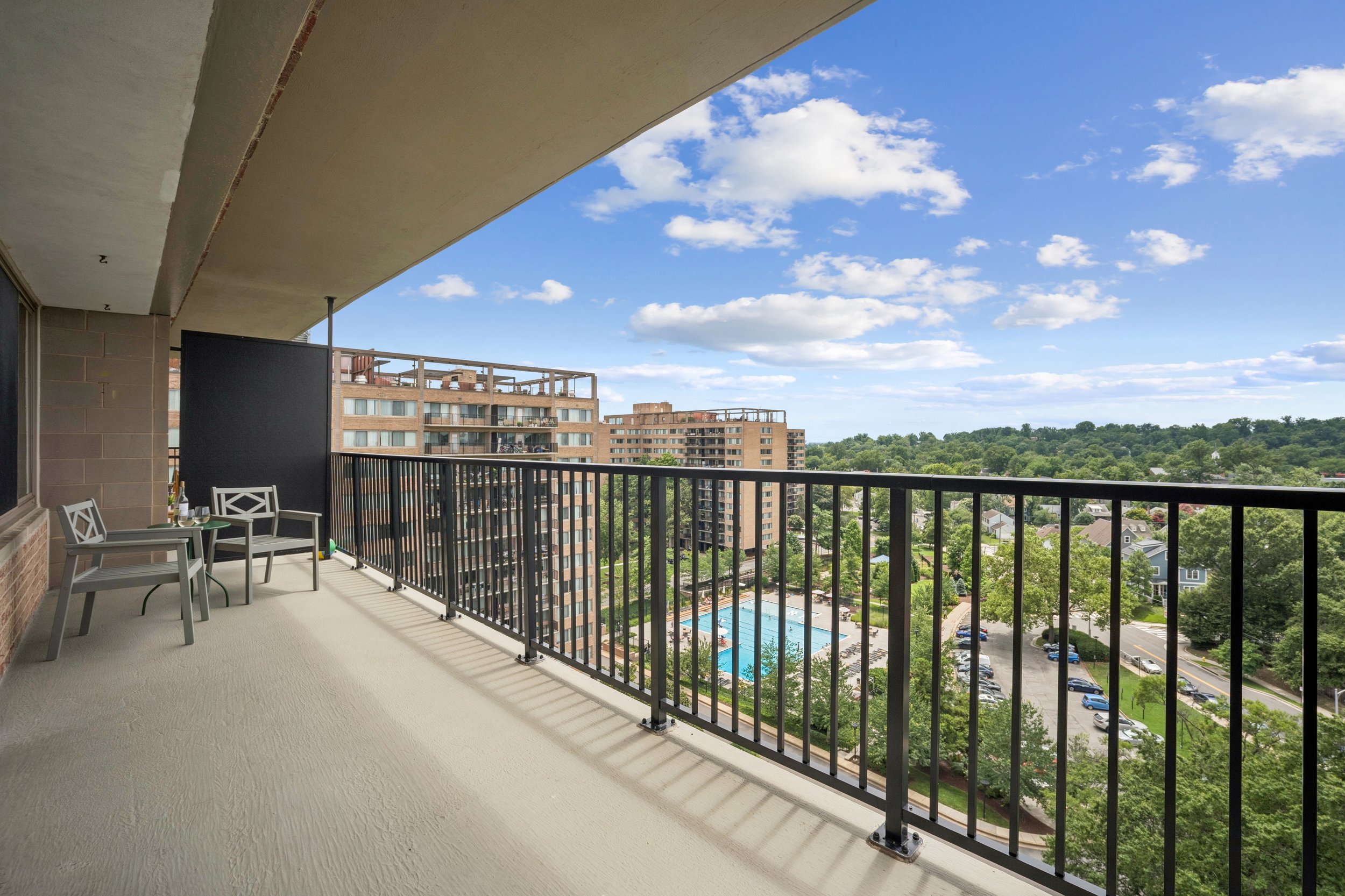 Balcony view overlooking a pool and apartment buildings, with trees and a blue sky with clouds in the background.