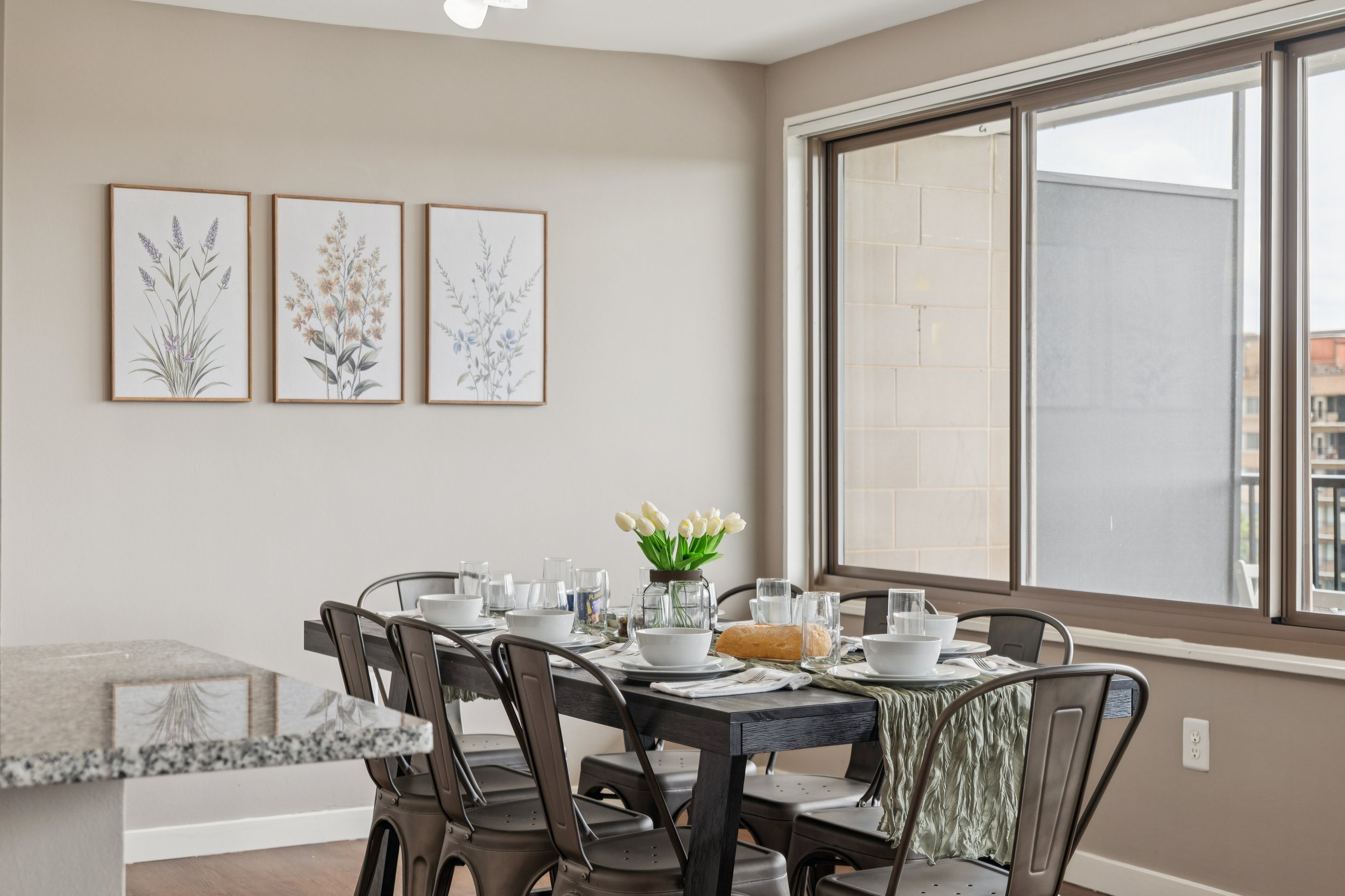 Dining table set with white dishes, glasses, a loaf of bread, and a vase with white tulips in a room with large windows and framed floral artwork on the wall.