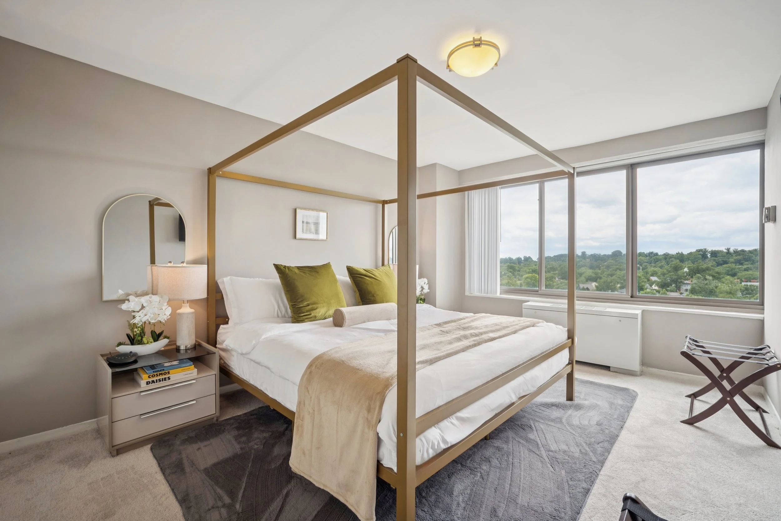A modern bedroom with a beige canopy bed, white linens, and green pillows. A nightstand with a lamp, books, and decorative items is beside the bed. Large windows show greenery outside, and a luggage rack is near the window.