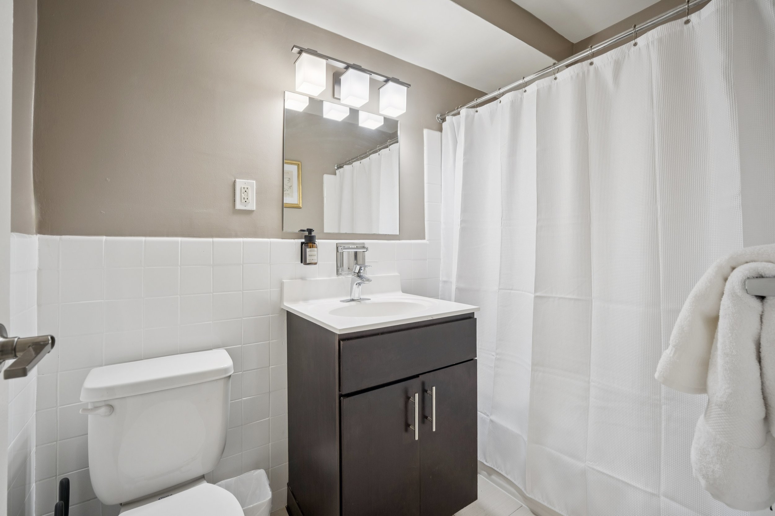 Modern bathroom with white tiled walls, a dark vanity with a sink, a mirror, a wall-mounted light fixture, a white toilet, a white shower curtain, and a towel on a hook.