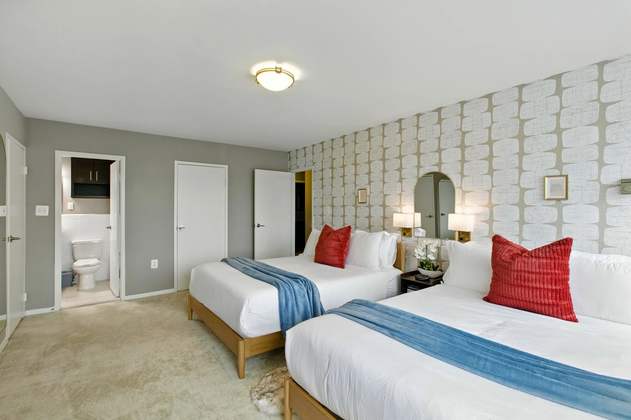 A hotel room with two beds, each with white linens, red pillows, and blue throws, a nightstand with a flower arrangement, and a patterned accent wall. There's a lamp, mirror, and framed artwork above the beds, and an open door leading to a bathroom.