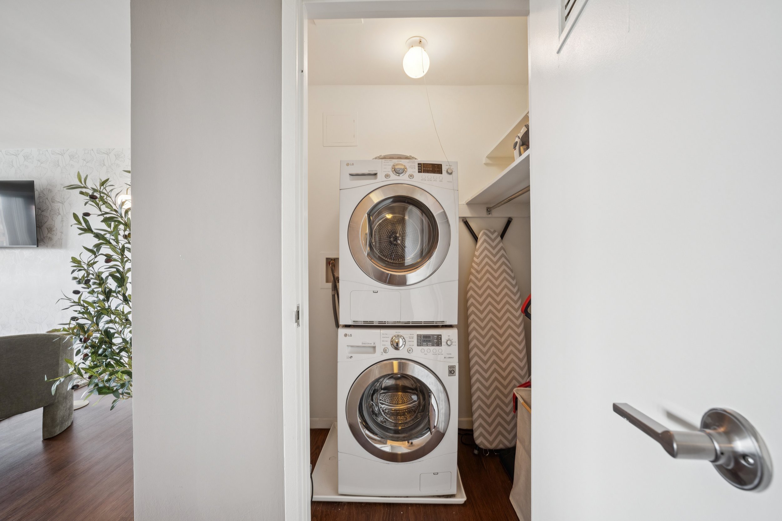 Stacked front-loading washing machine and dryer in a laundry closet, with an iron and ironing board beside them.