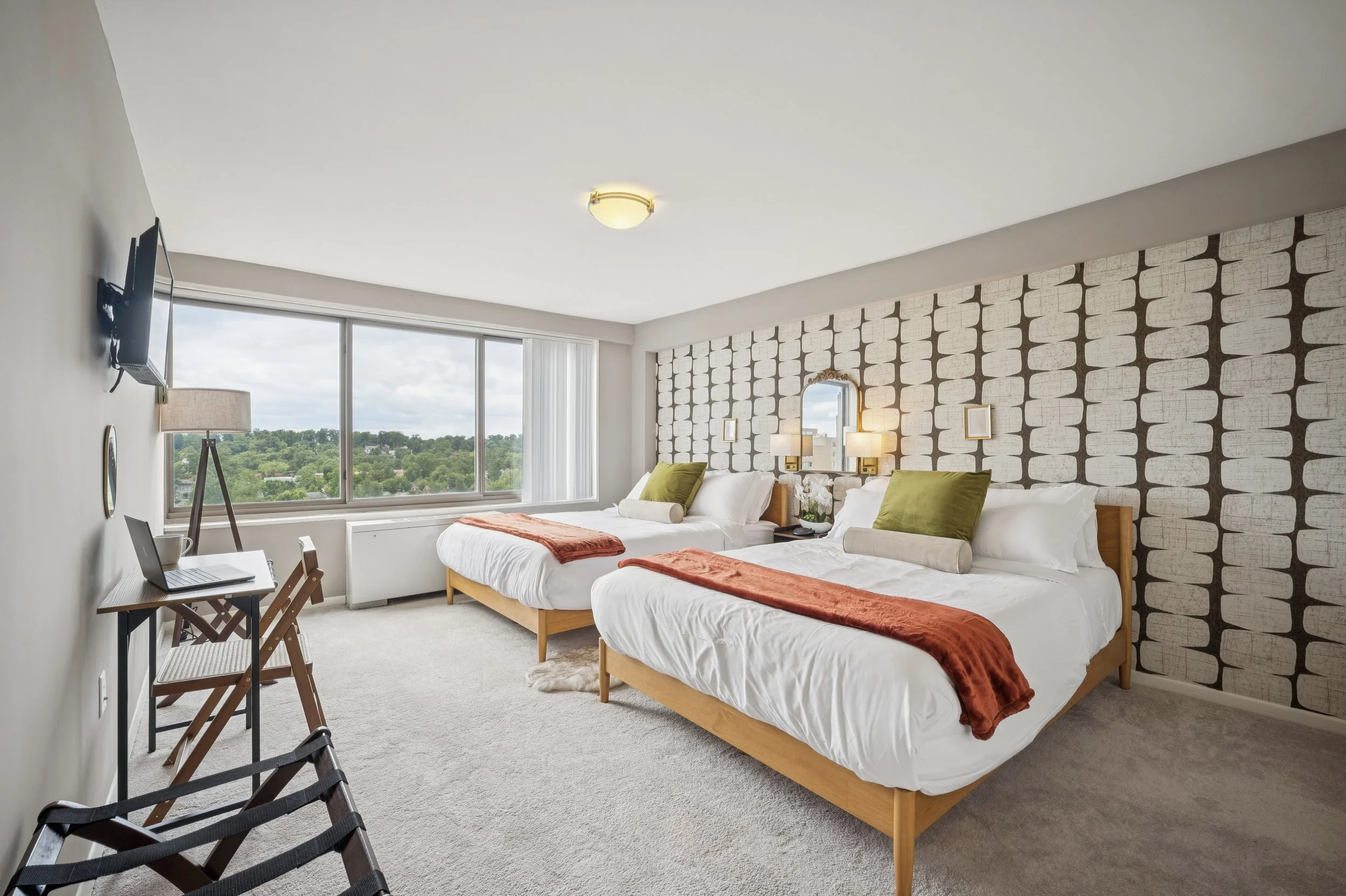 A hotel room featuring two beds with green and white pillows, a large window with a city view, a dresser with a lamp, a wall-mounted TV, and modern decor.