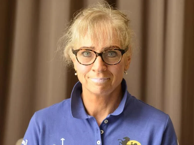 A woman with blonde hair, glasses, and pearl earrings wearing a blue polo shirt with a logo on the chest, standing in front of a neutral curtain background.