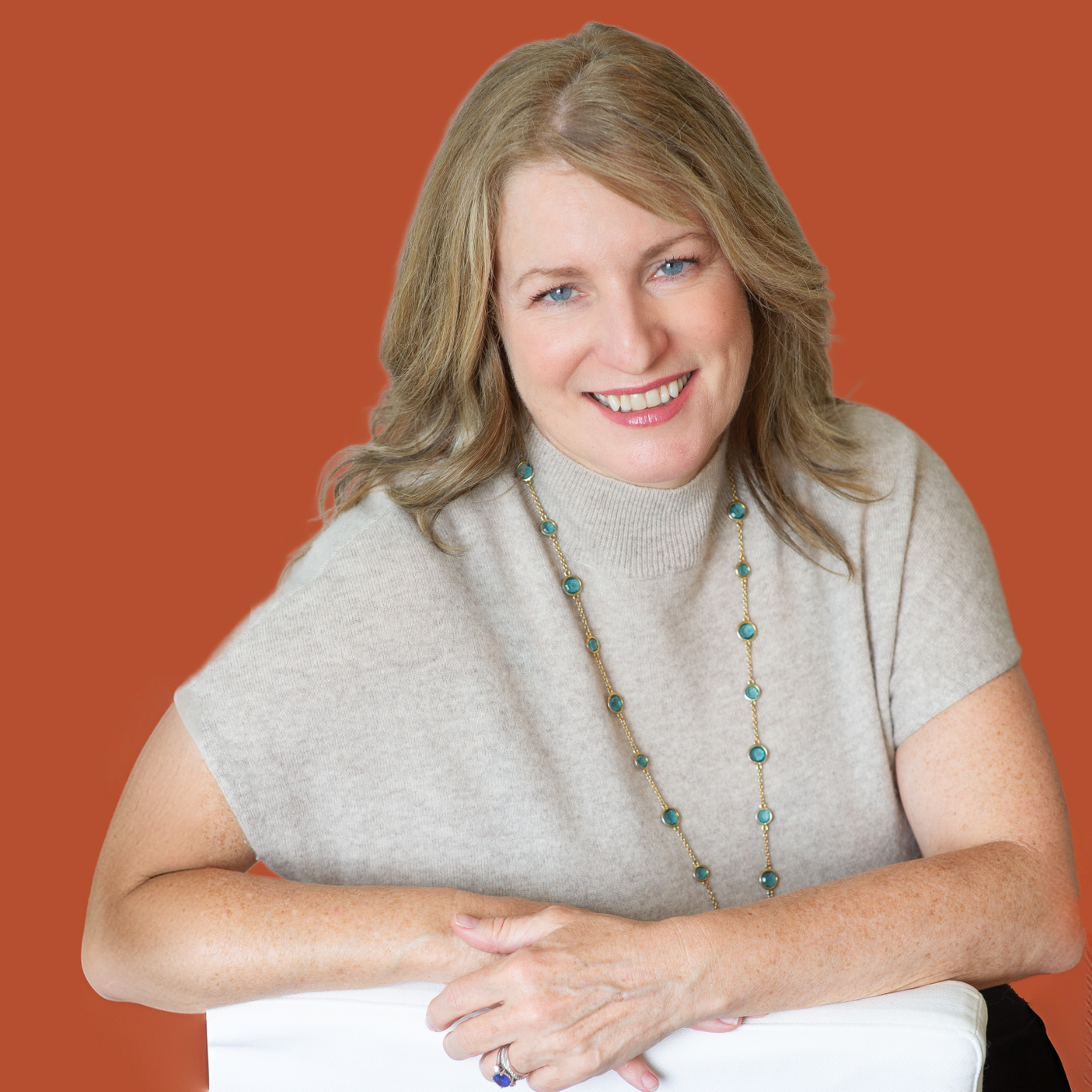 In front of a marigold background sits a white woman with blonde hair and blue eyes. She is smiling and wearing a biege sweater with a long gold and green necklace over top.