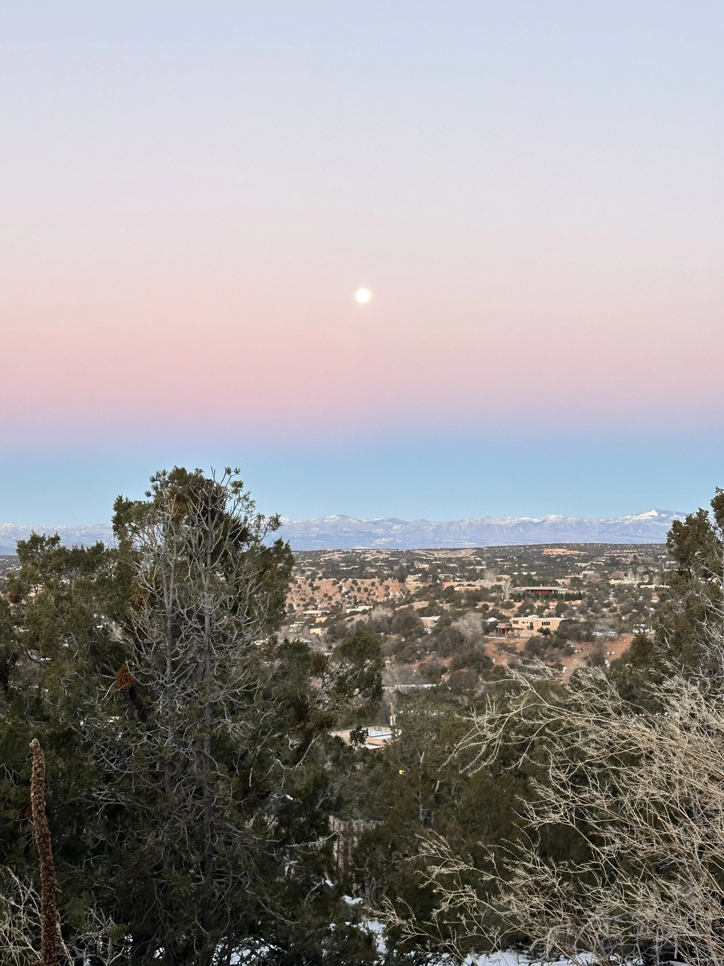 A with a pastel pink and blue sky sunset in Santa Fe, New Mexico, a visible full moon, distant snow-capped mountains, and trees in the foreground.