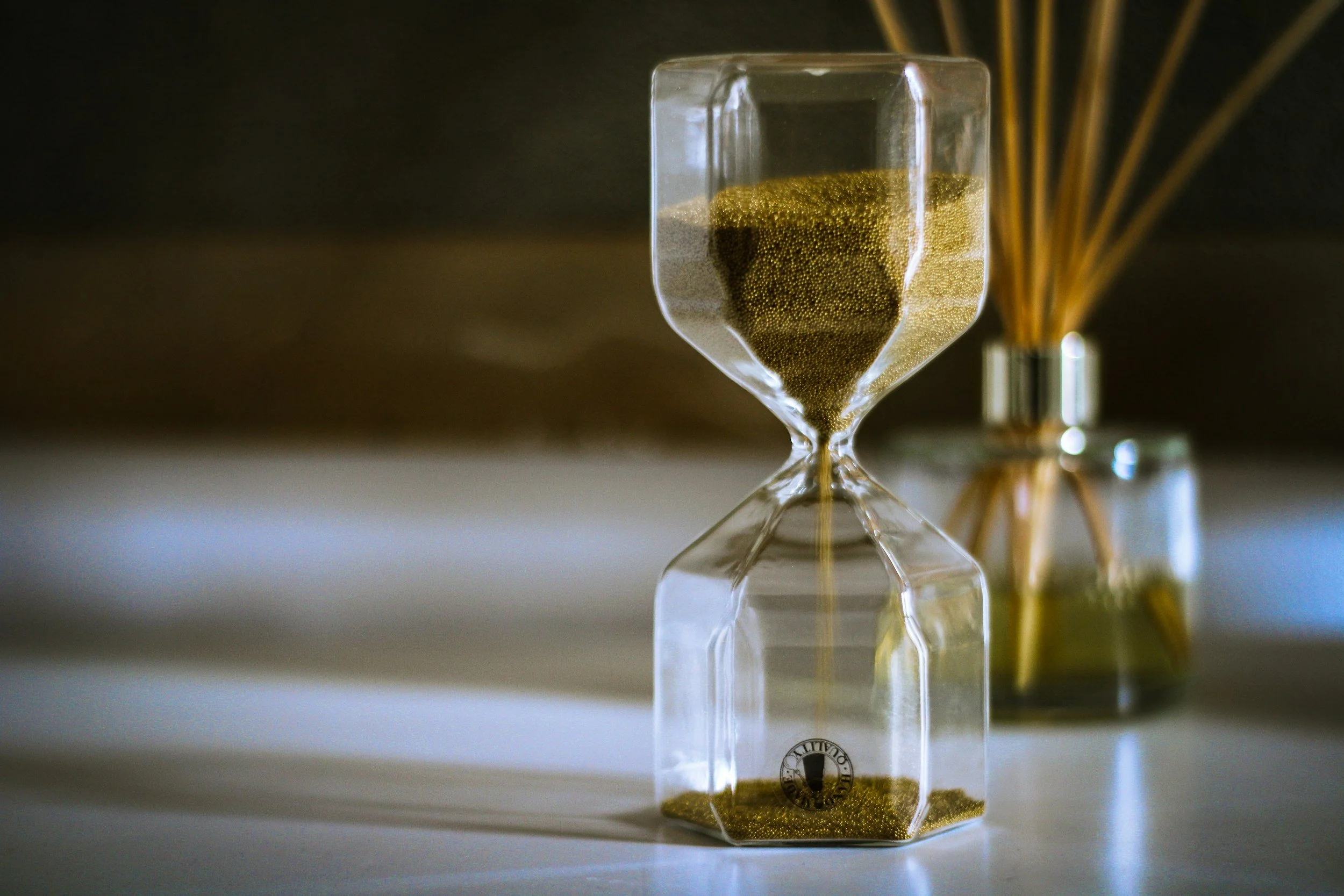 An hourglass with golden sand and a small jar of matching sand behind it, placed on a light-colored surface.