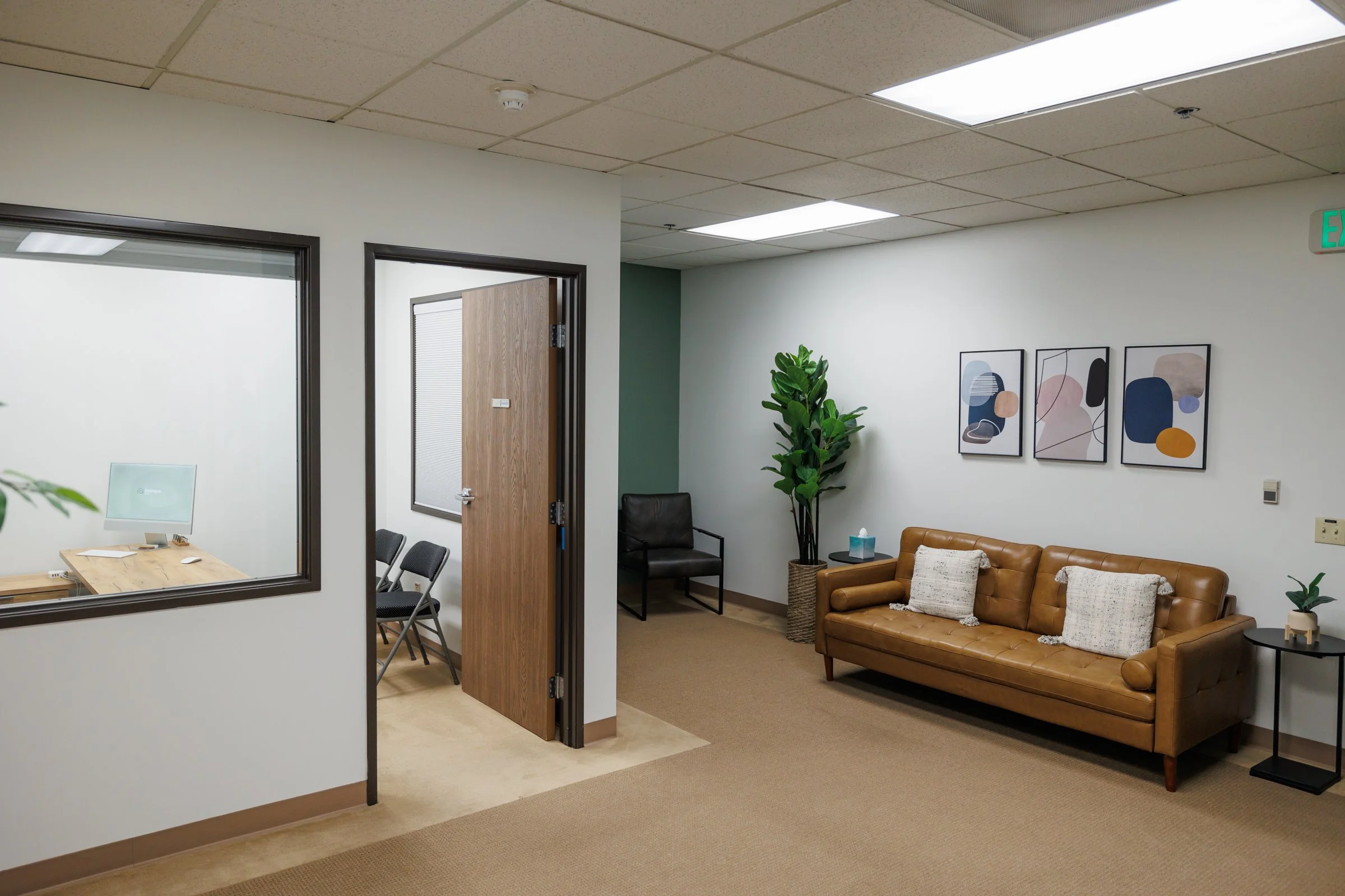 Office waiting area with a brown leather couch, a small side table with a plant, a black armchair, a tall potted plant, and three framed abstract art pieces on the wall.