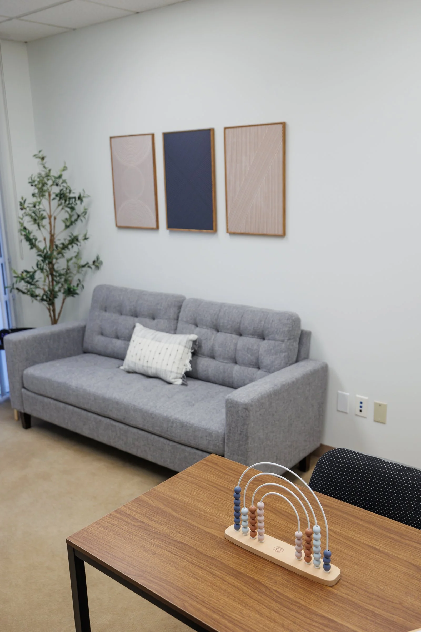 A cozy room with a gray upholstered sofa, a decorative pillow, a small wooden table with a baby abacus, framed wall art, a potted plant, and electrical outlets on a plain white wall.