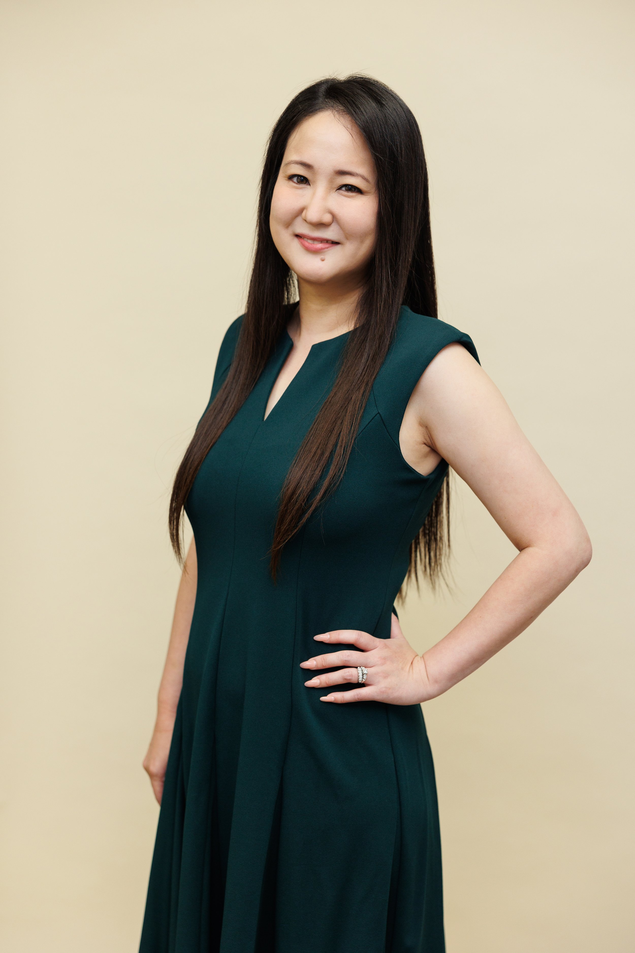 A woman with long dark hair smiling and winking, wearing a white quilted dress with a belt, standing against a neutral background.