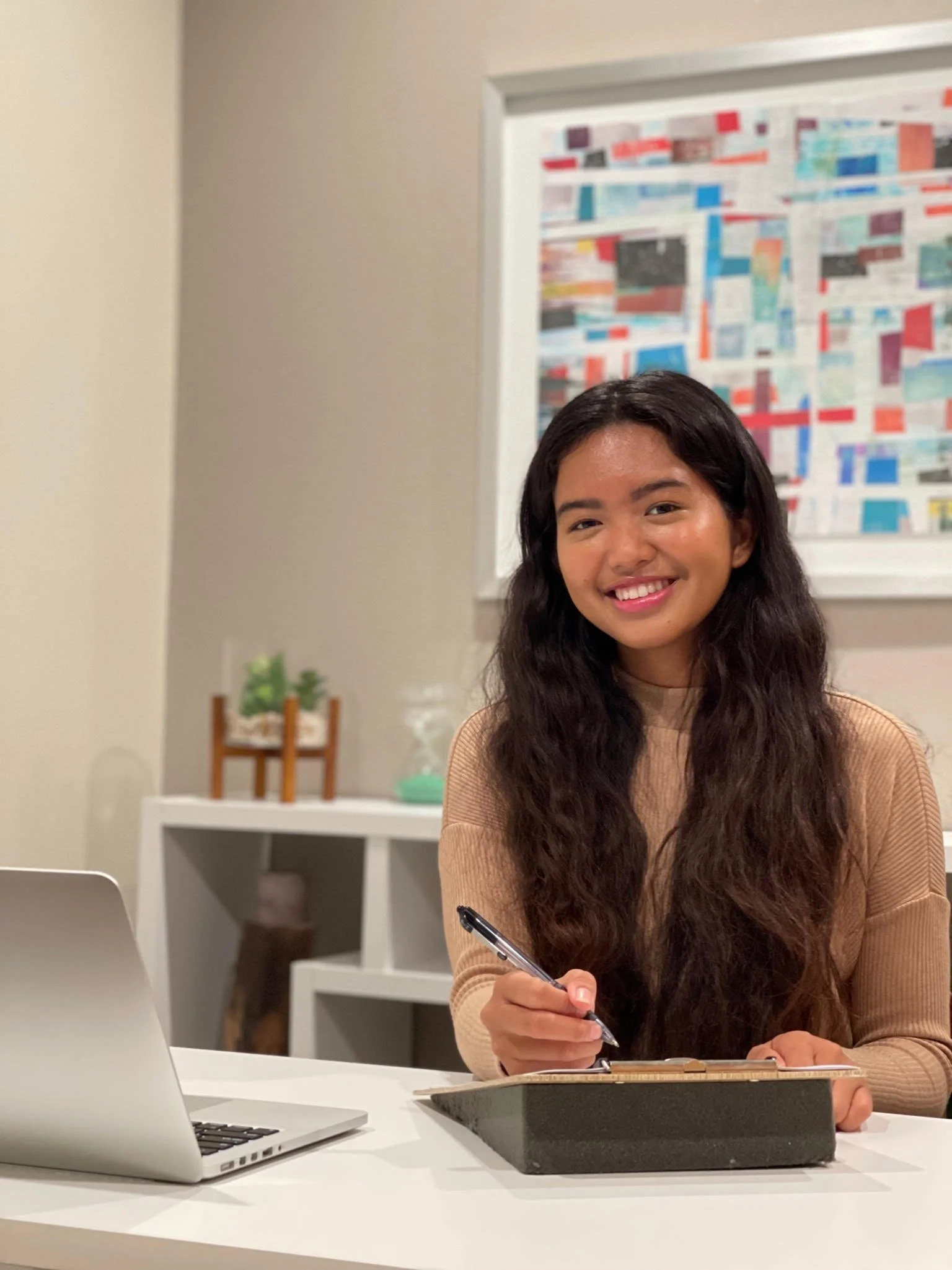 Young woman with long dark hair smiling at camera while sitting at a white desk with a laptop and writing in a notebook, in a modern office space.