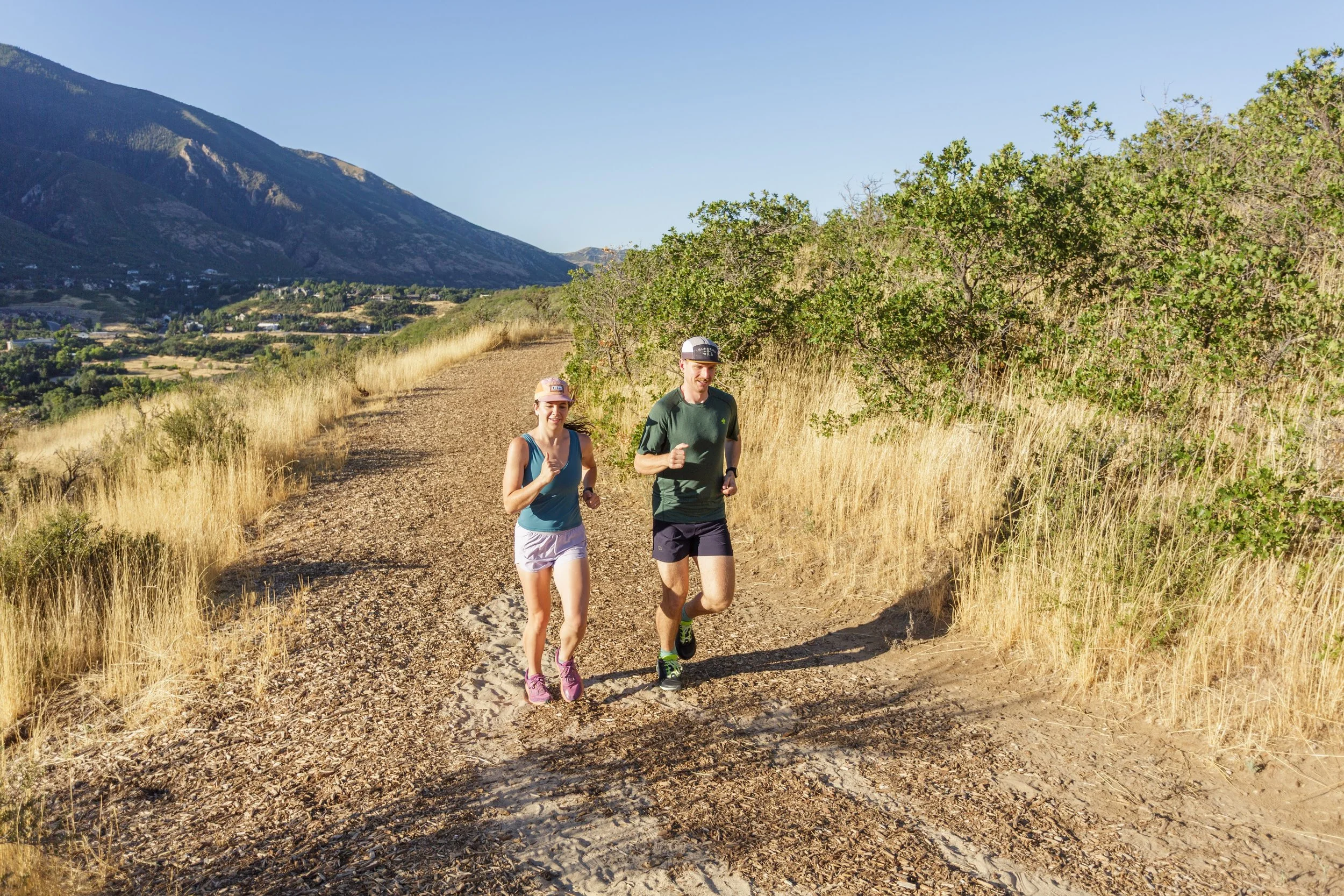 Two people, a woman and a man, jogging on a dirt trail through a grassy hillside with trees, mountains in the background, and a clear blue sky.