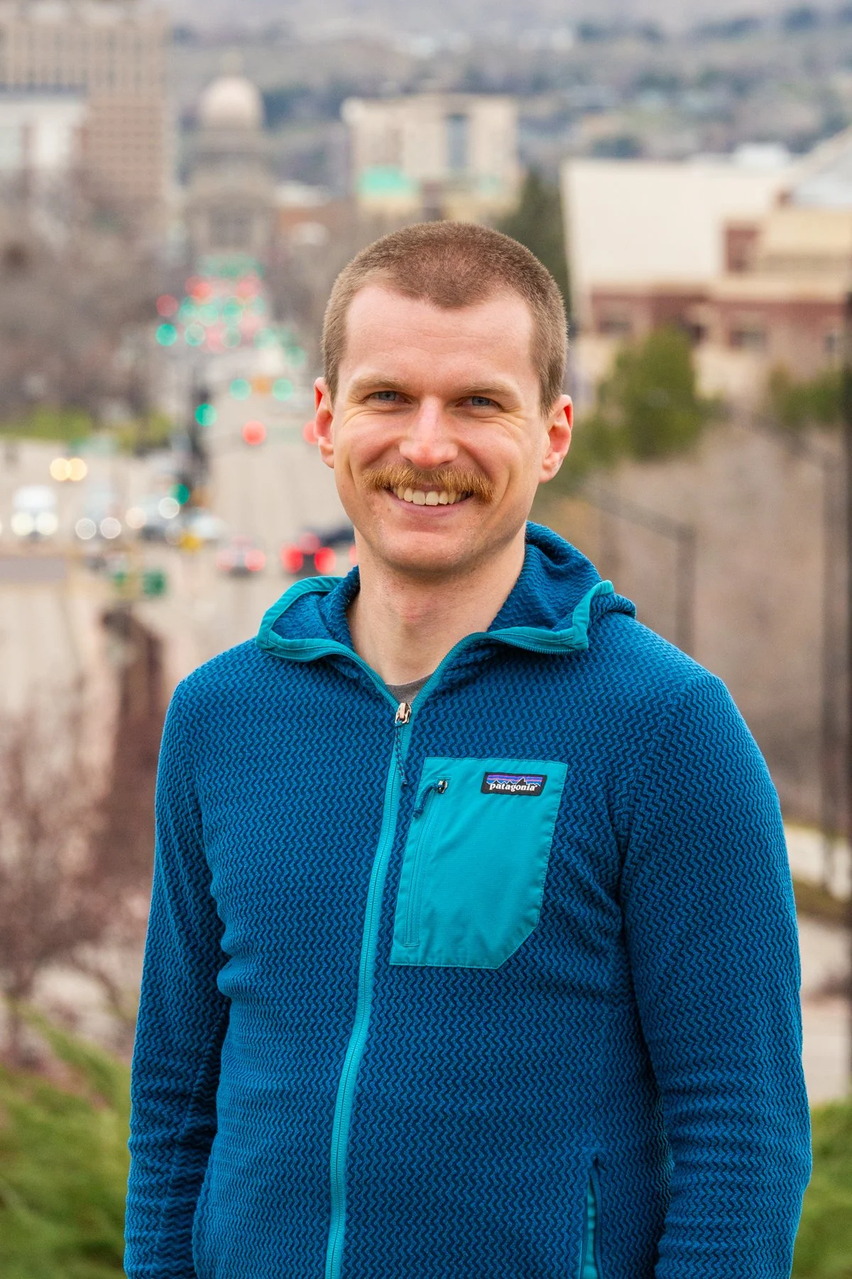 A smiling man with short hair, a mustache, and blue eyes, wearing a blue Patagonia fleece jacket, standing outdoors on a city street with blurred buildings and cars in the background.