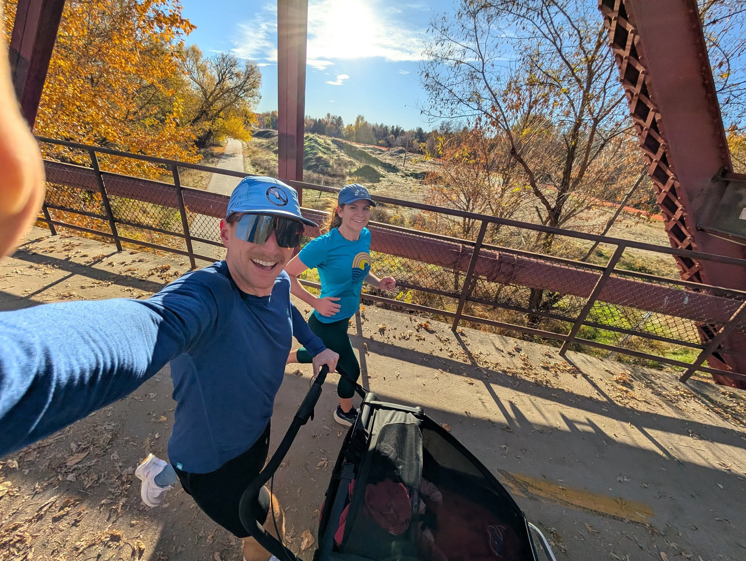 Two people smiling and jogging on a bridge during autumn, with one pushing a stroller, surrounded by trees with colorful fall leaves, under a sunny blue sky.