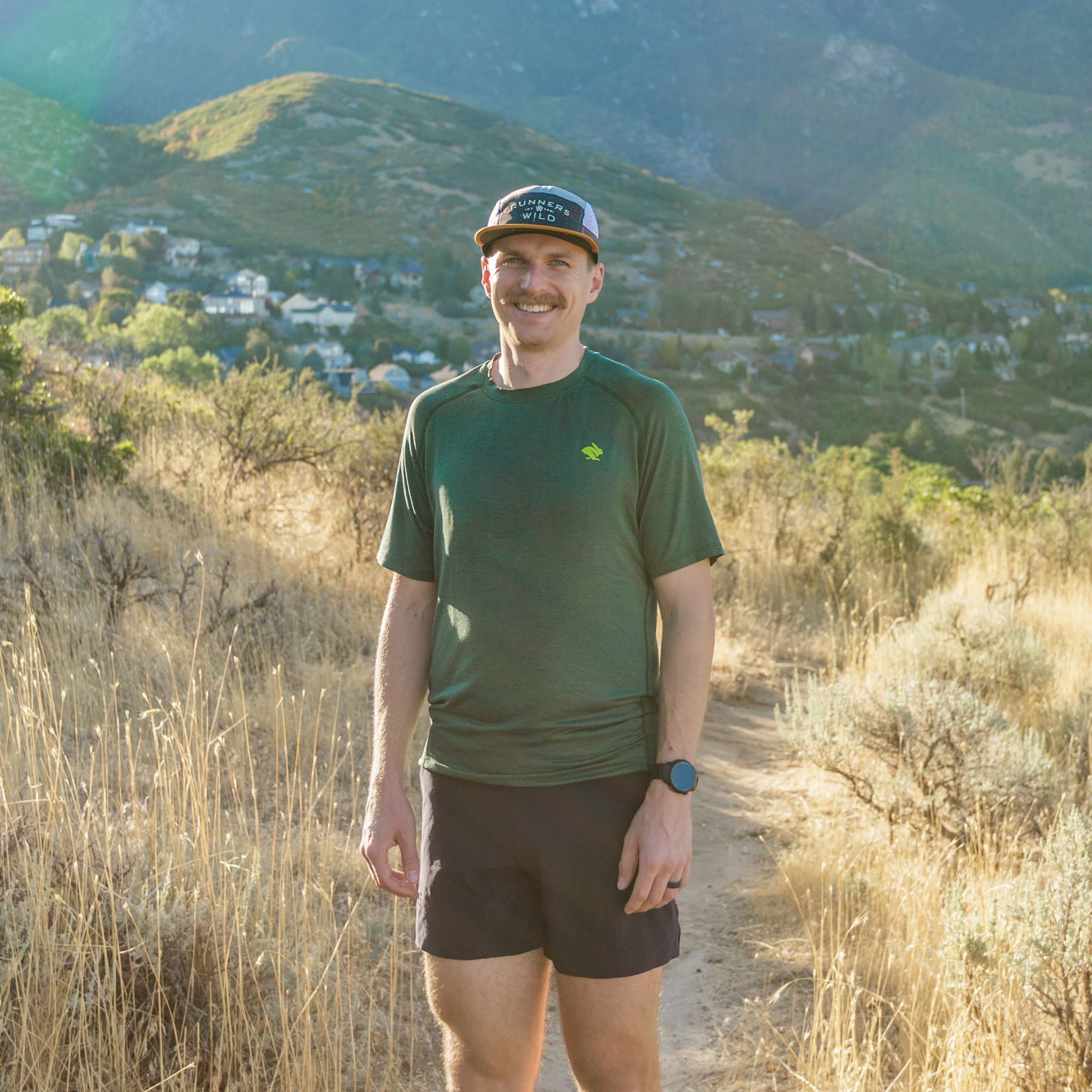 A man with a hat, mustache, wearing a green shirt and black shorts, smiling outdoors on a trail with hills and houses in the background.