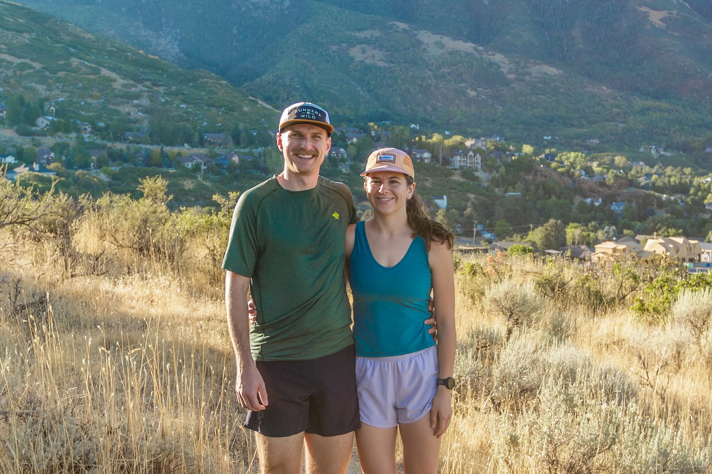 A young man and woman smiling and standing together outdoors on a grassy hillside with trees, houses, and mountains in the background on a sunny day.