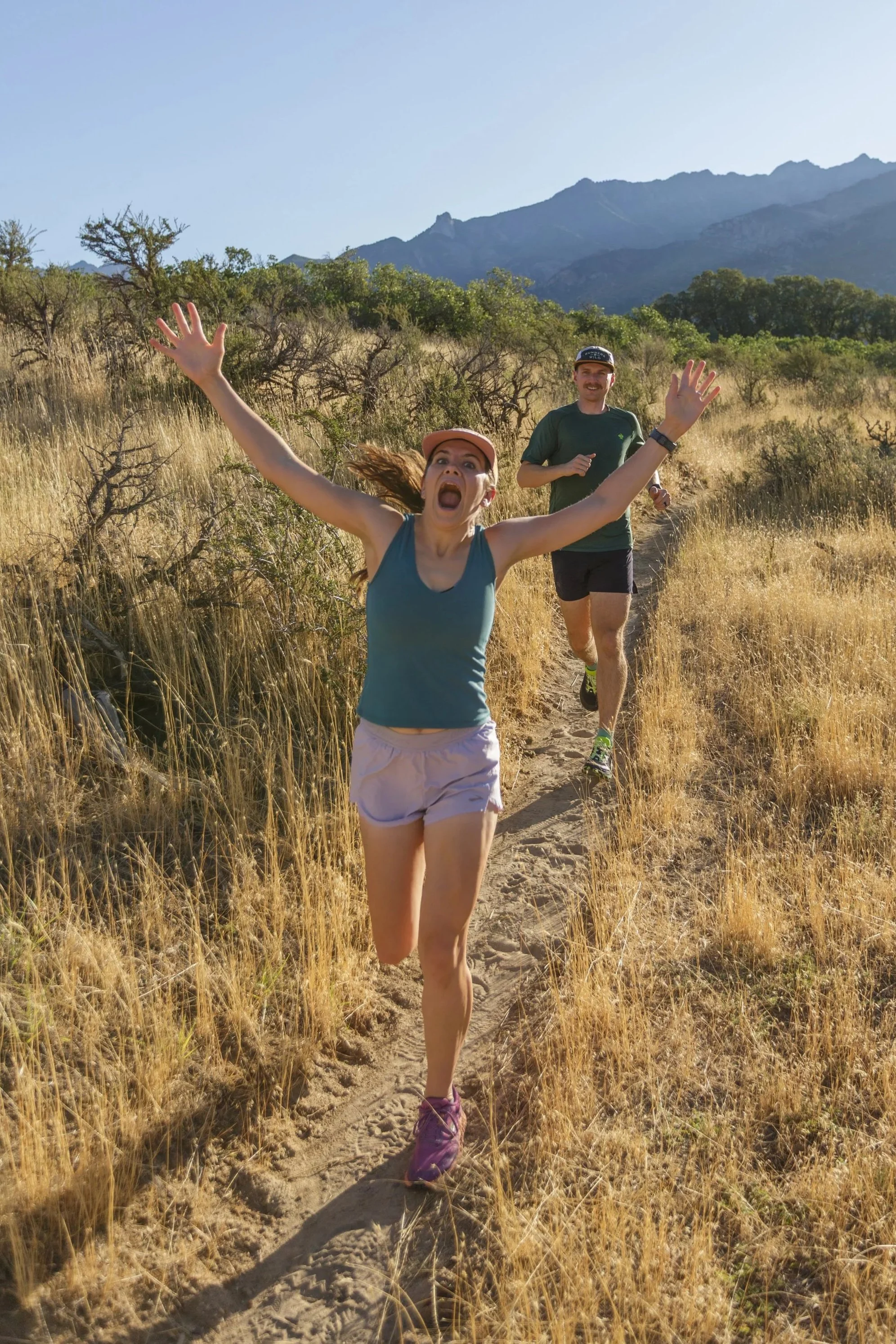 A woman and a man running on a dirt trail through a grassy field with trees and mountains in the background on a sunny day.
