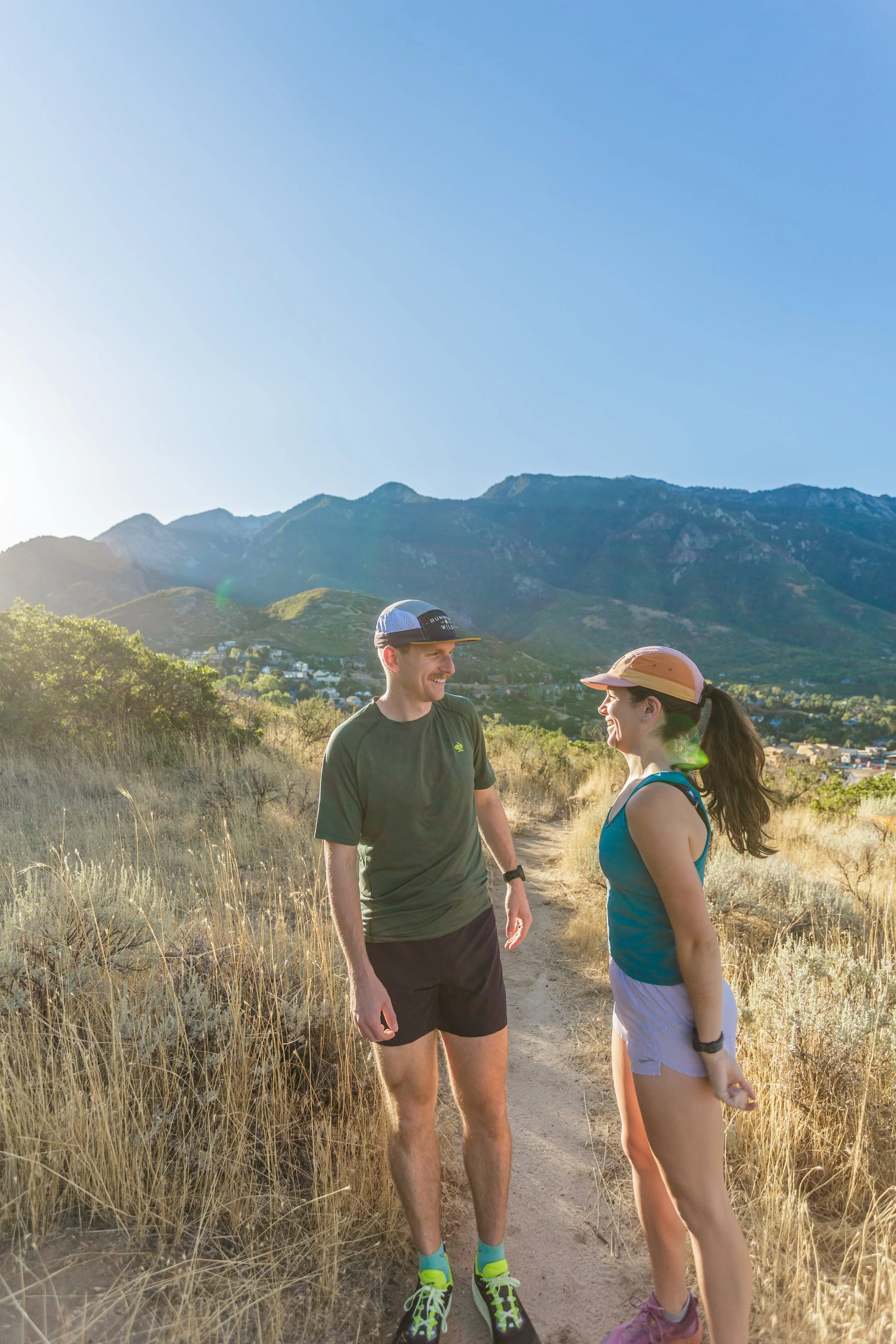 A man and a woman stand on a dirt trail amidst tall grass, with mountains in the background under a clear blue sky. They are engaged in conversation, smiling at each other, dressed in athletic wear, and appear to be enjoying a hike or outdoor activity.