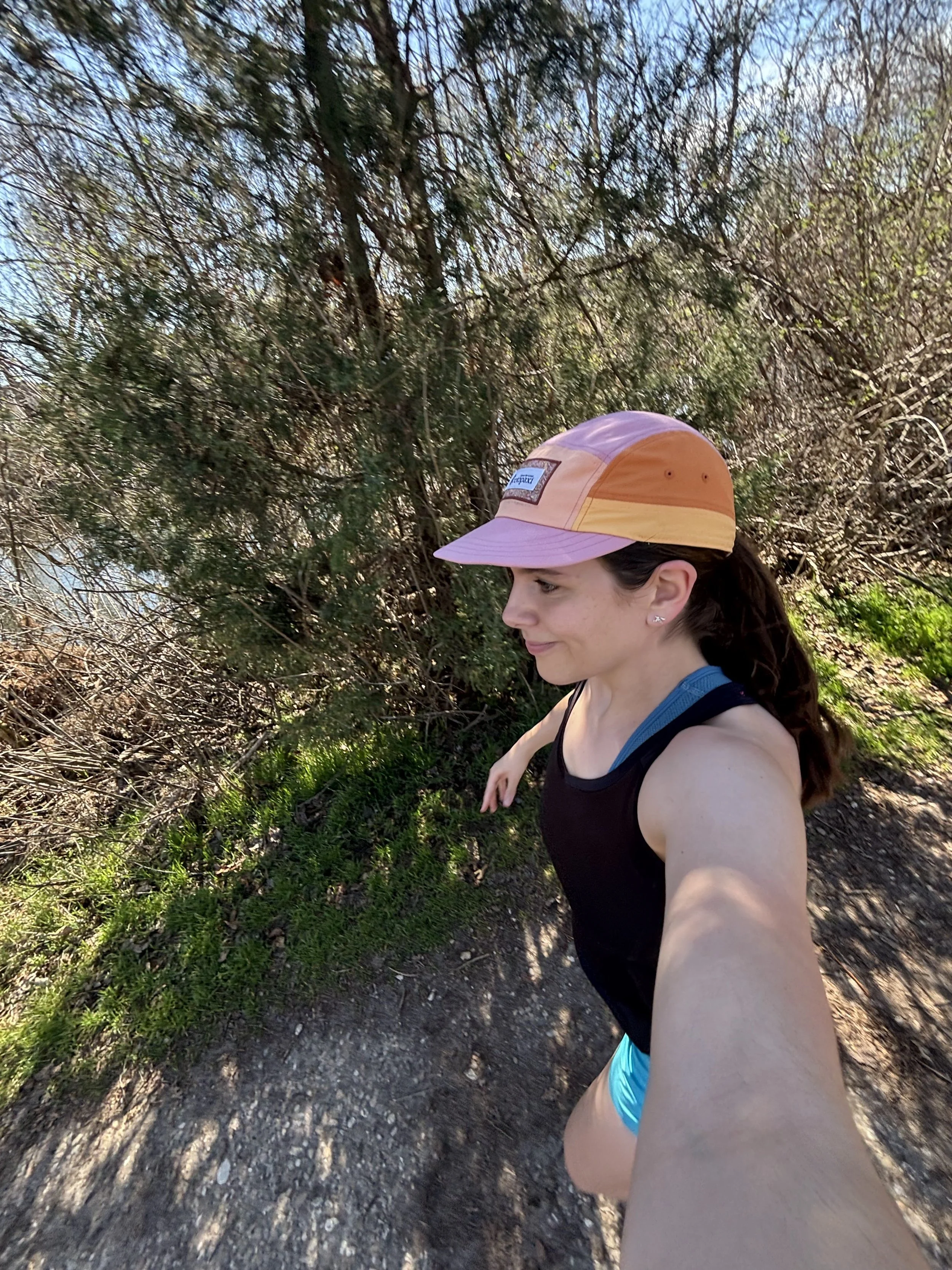 A woman running on a trail surrounded by bushes and trees, wearing a pink and orange cap, a black tank top, and blue shorts, taking a selfie during the daytime.