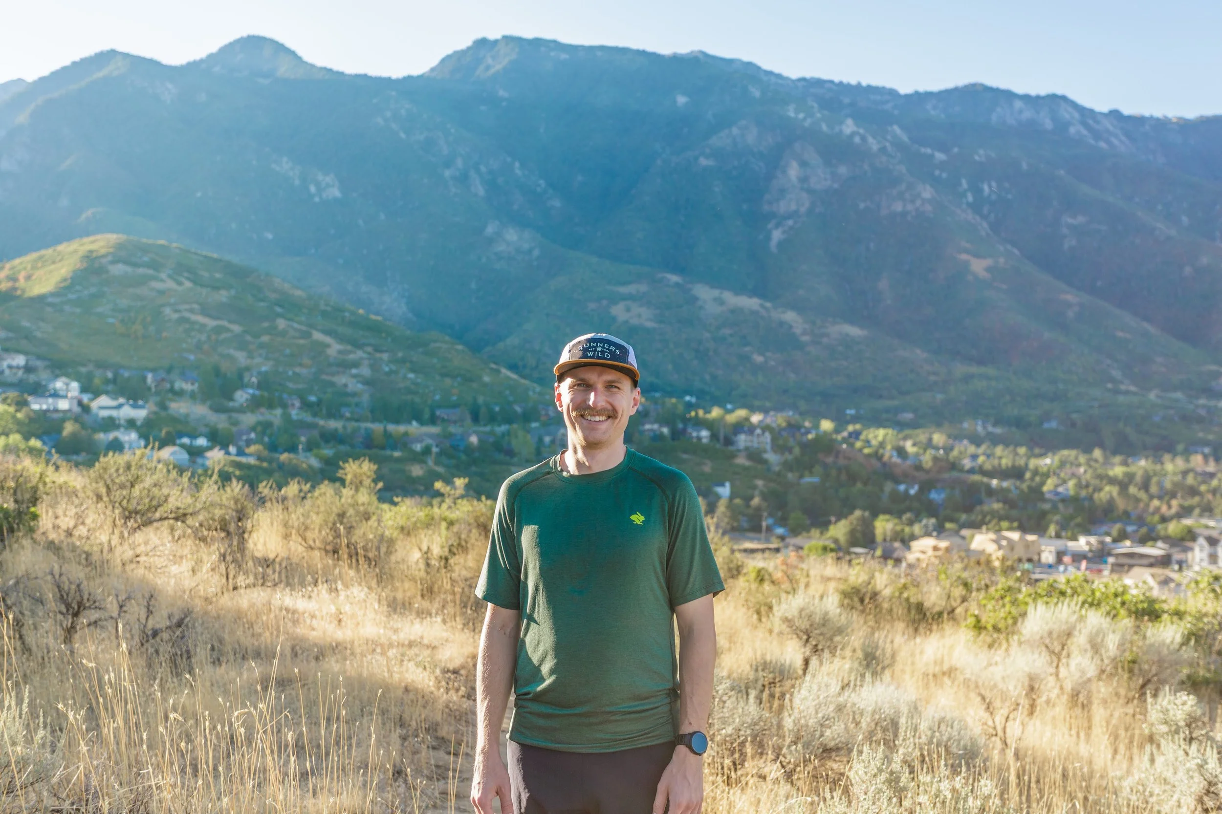 A smiling man wearing a green long sleeve shirt, a watch, and a black cap that says 'RUNNERS WILD' stands outdoors on a trail with dry grass. Behind him, there are green hills and a mountainous landscape, with a town visible at the base of the hills.
