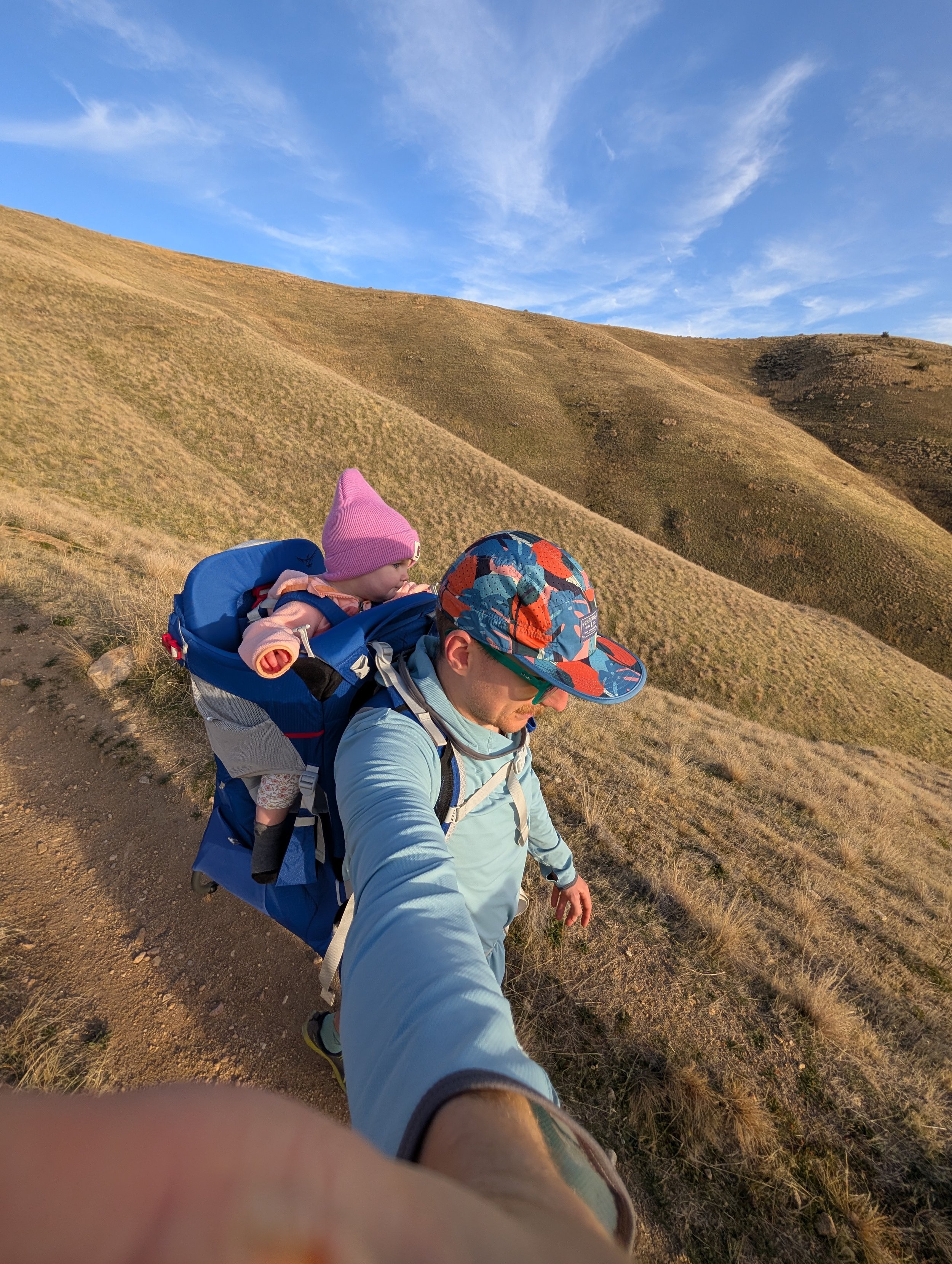 A man hiking with a child in a backpack carrier on a trail through grassy hills with a blue sky.
