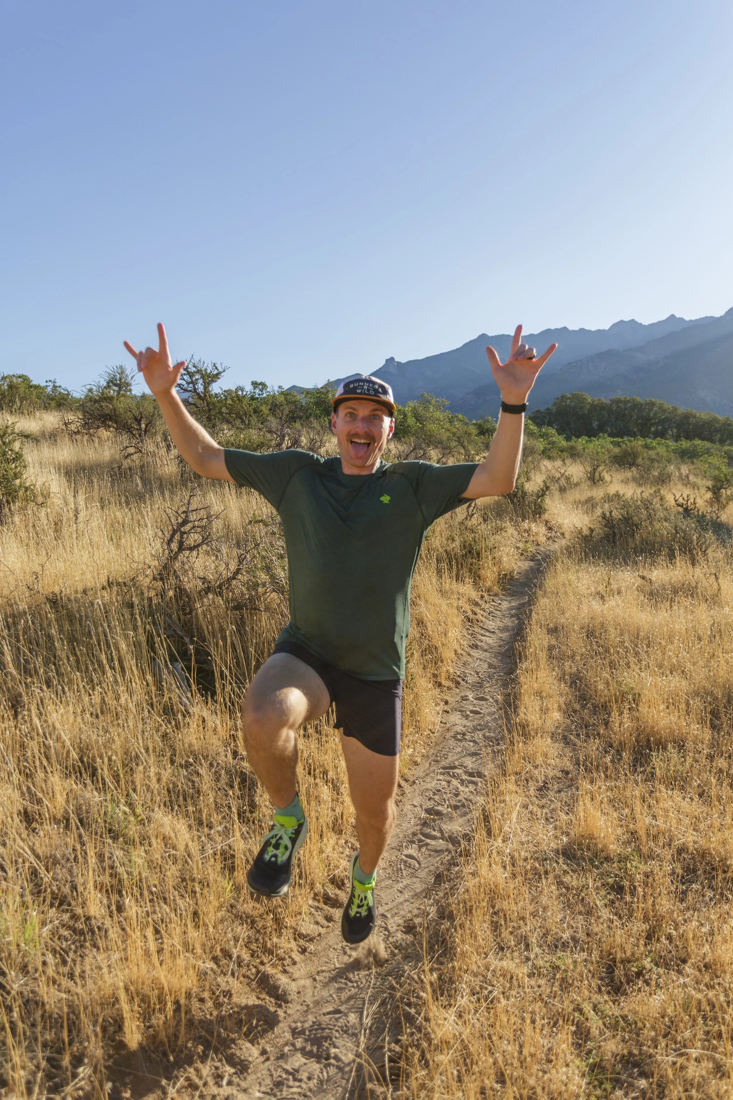 A man wearing a gray shirt, black shorts, and running shoes is jumping on a narrow dirt trail in a grassy field with shrubs and mountains in the background. He is smiling and making a hand gesture with both hands.