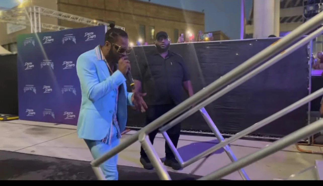 A man in a light blue suit with sunglasses holding a microphone and performing at an outdoor event at night.