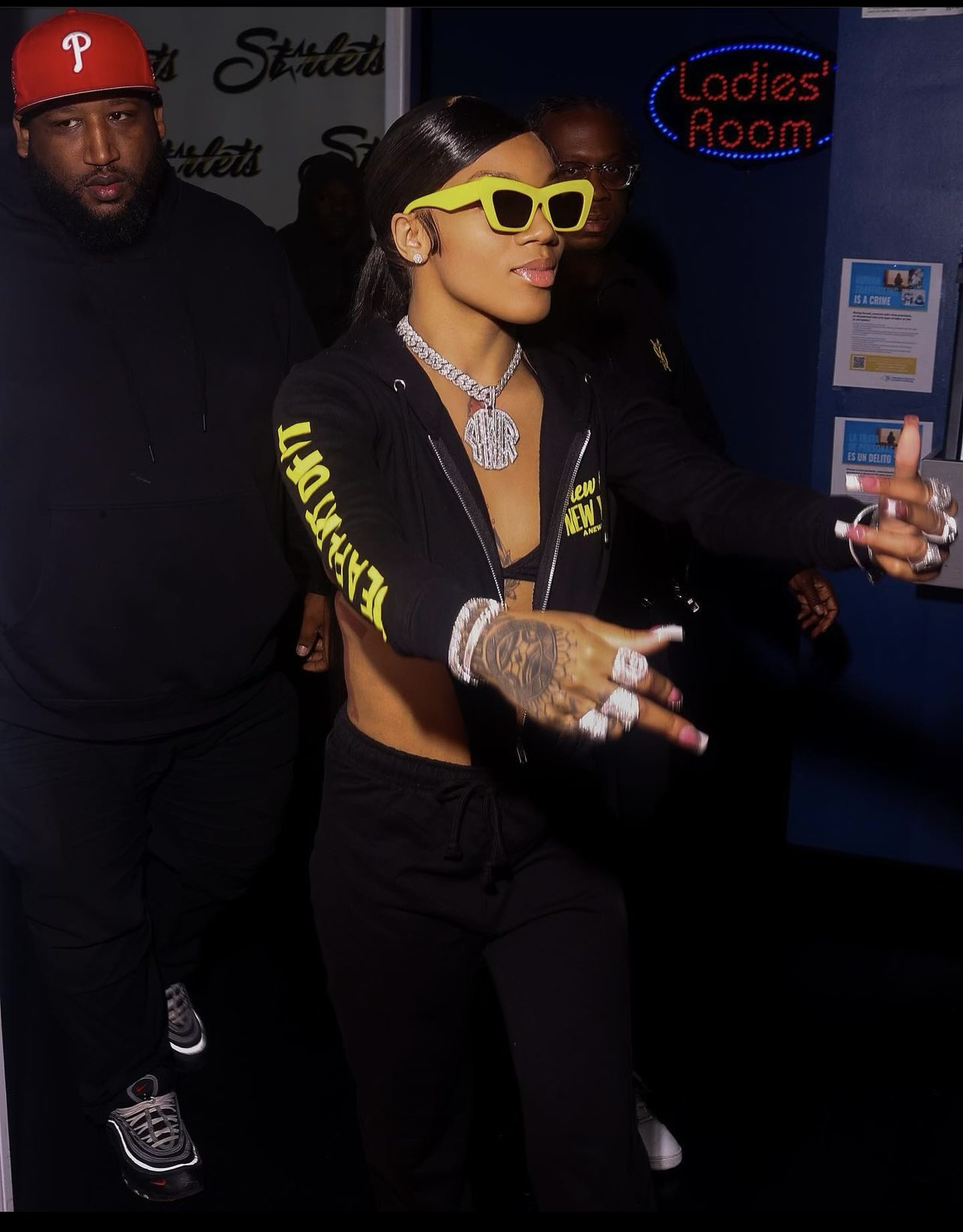 A woman wearing yellow sunglasses and jewelry is taking a selfie with her phone, surrounded by security personnel, in an indoor setting with a sign that says 'Ladies Room'.