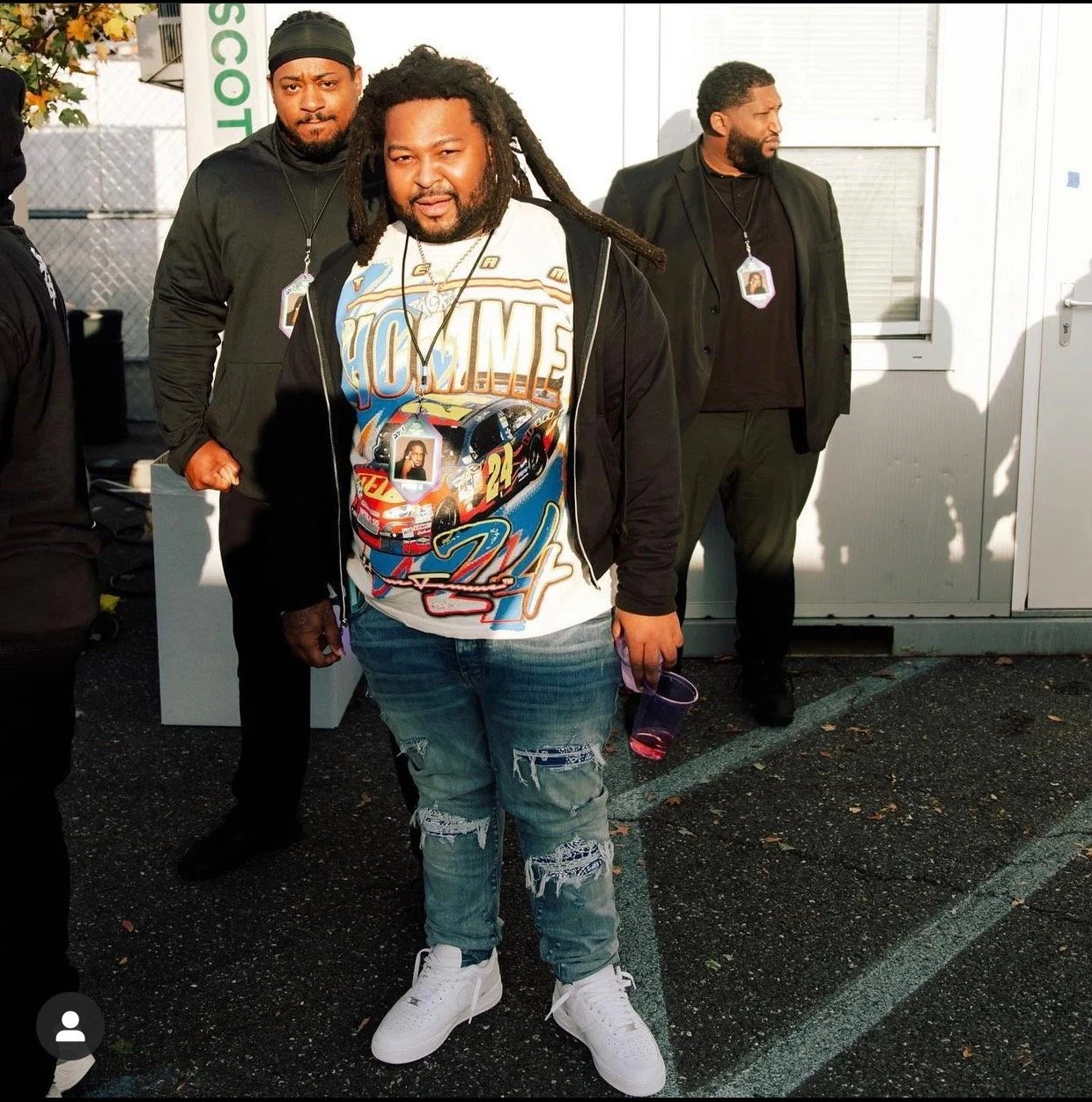 A man with dreadlocks wearing a racing-themed t-shirt, black hoodie, and distressed jeans, holding a pink drink, standing outside with two men in black suits and security badges behind him.