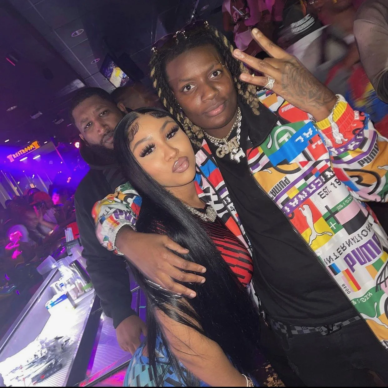 Three people at a nightclub; man with dreadlocks and colorful jacket making peace sign, woman with long black hair and makeup in front, man in black behind, purple lighting, crowded dance floor.