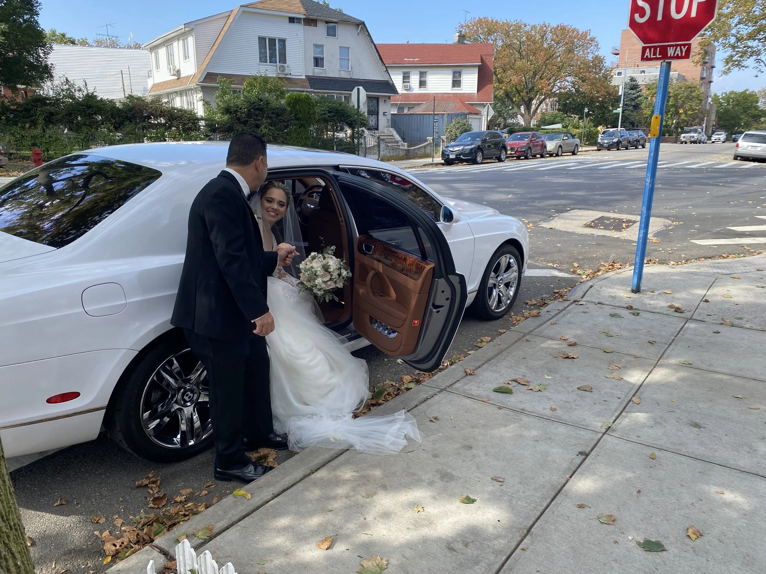 Bride with a bouquet getting into a white luxury car with a man helping her, on a city street with houses in the background, trees with fall leaves, and a stop sign.