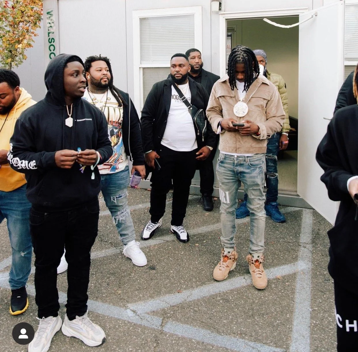 Group of men standing outside a building, some are looking at their phones, wearing casual streetwear including hoodies, jackets, and sneakers.