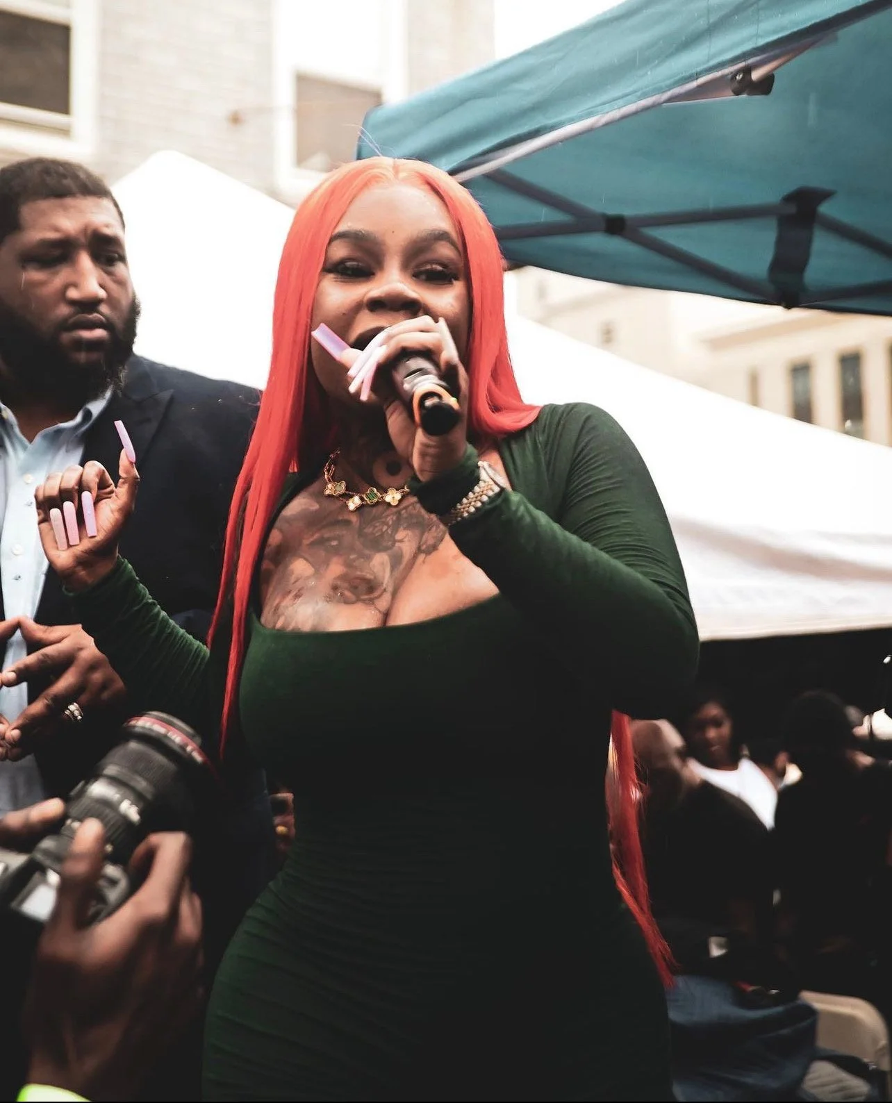 A woman with long red hair wearing a dark green dress holding a microphone at an outdoor event, with a man standing nearby and a group of people in the background.