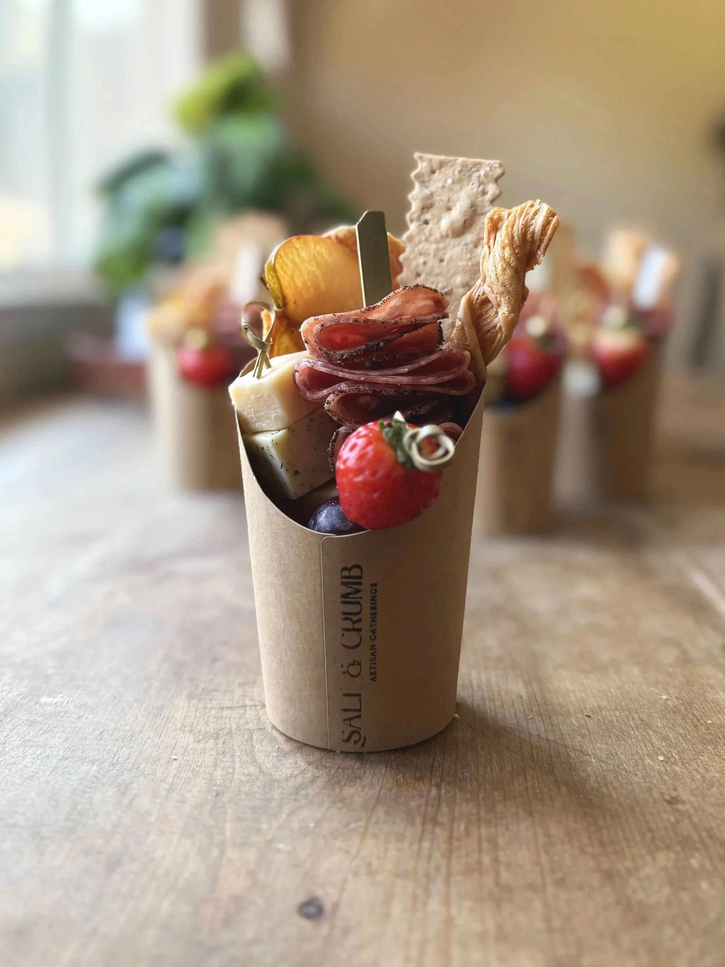 A charcuterie cup filled with cheese, sliced meats, strawberries, blueberries, and crackers on a wooden table.