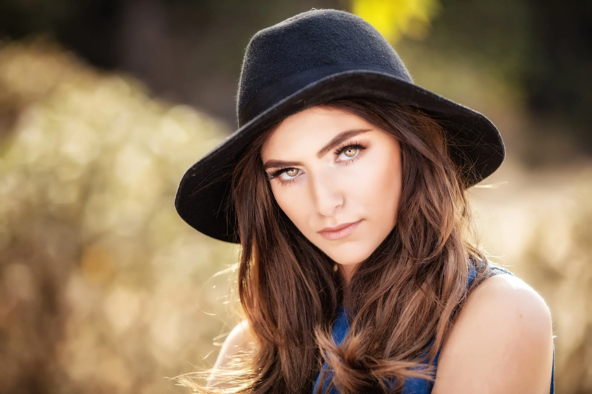 A young woman with long brown hair, green eyes, wearing a black wide-brimmed hat and a blue top, looking at the camera outdoors.