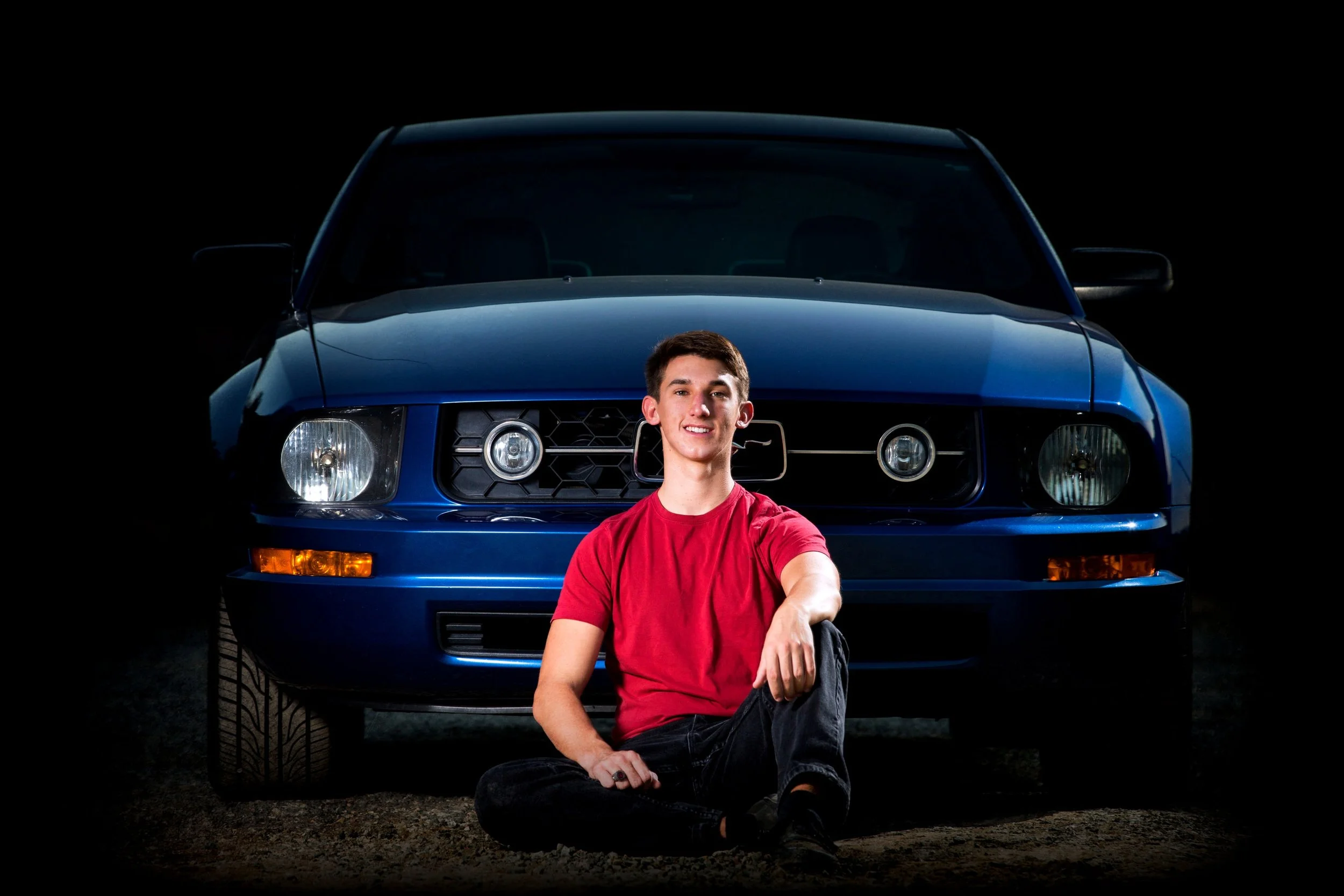 Young man sitting on the ground in front of a blue classic car, with a dark background. The man is smiling and wearing a red t-shirt and black jeans.