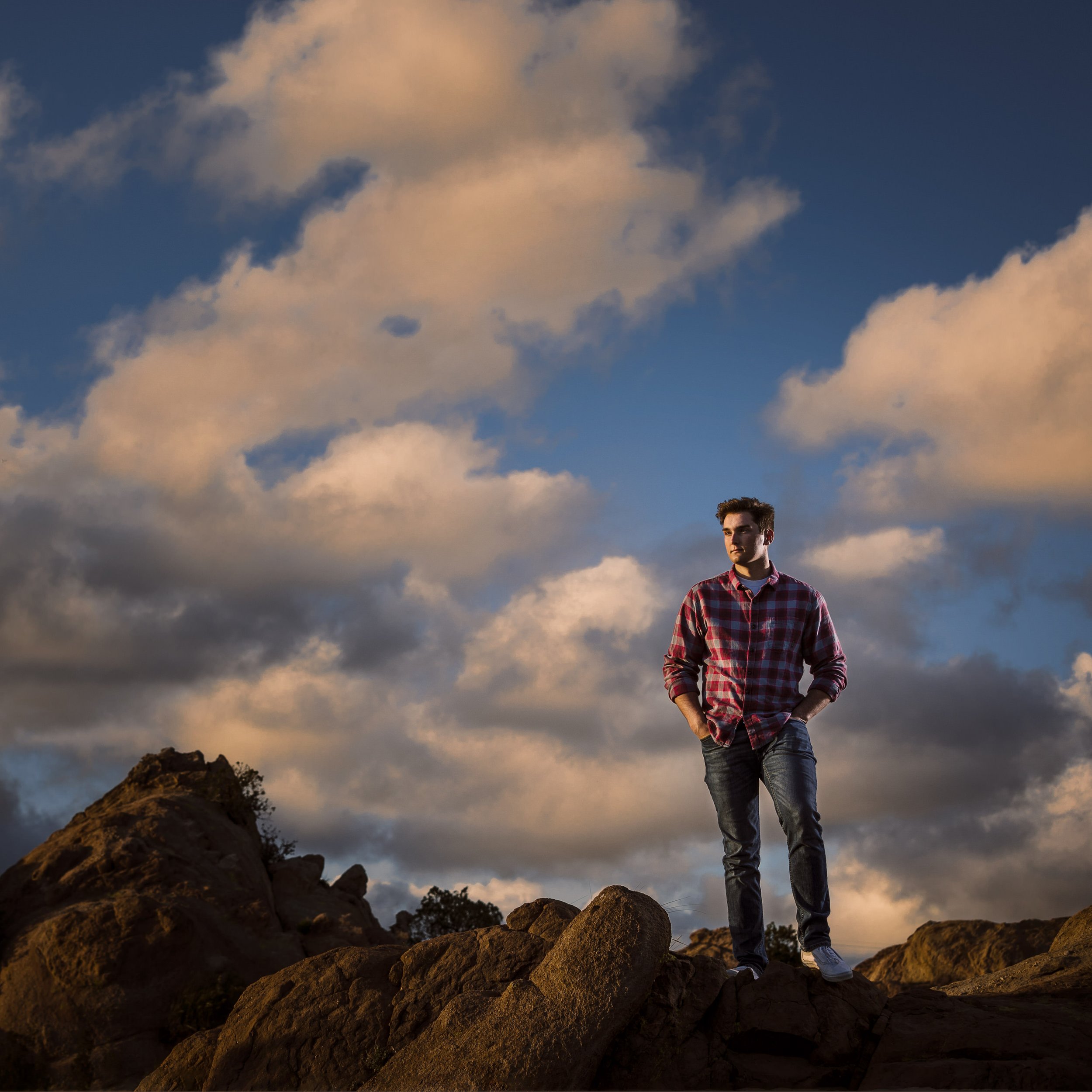 A young man in a red plaid shirt and jeans standing on rocks outdoors during sunset with a sky full of clouds in the background.