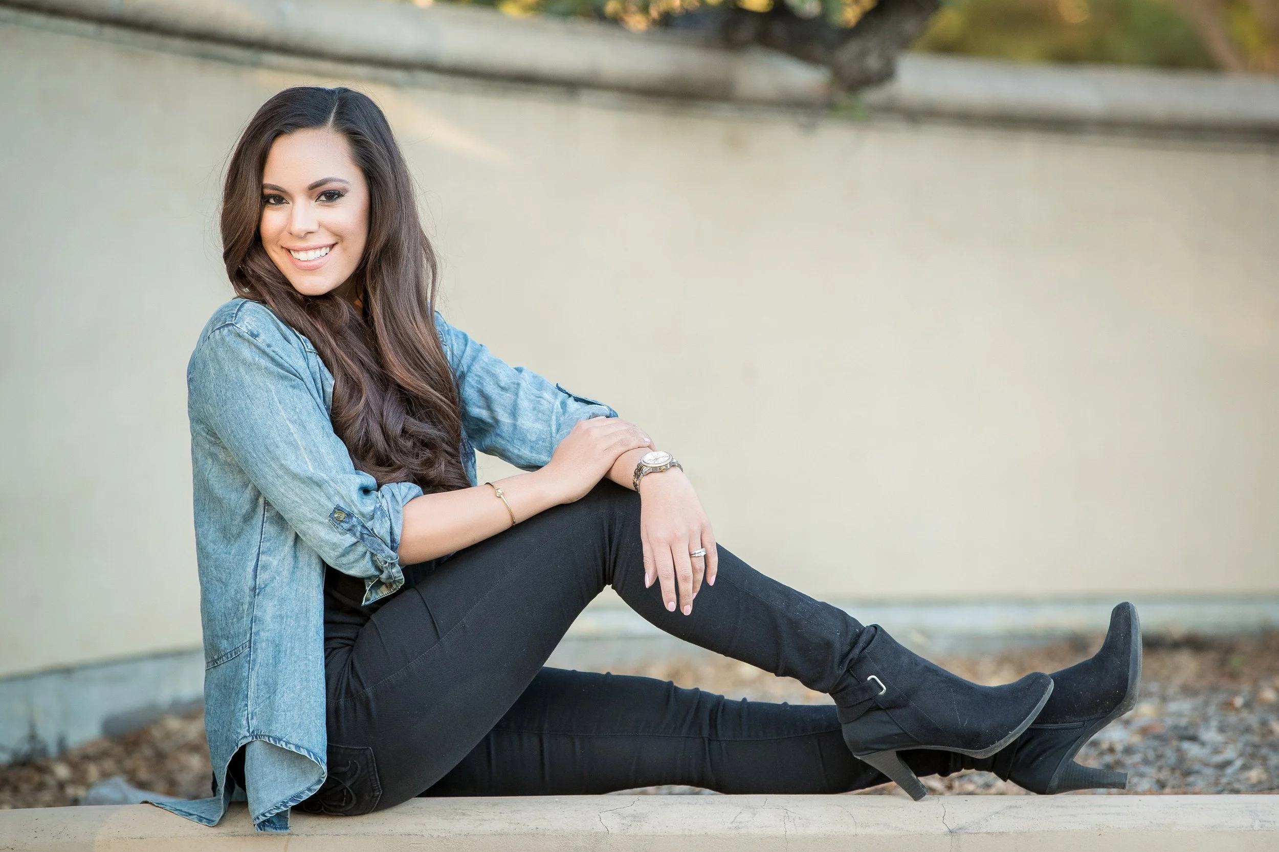 A young woman with long brown hair is sitting outdoors, smiling, wearing a light blue denim jacket, black pants, and black heeled boots. She has jewelry including a watch, rings, and a bracelet.