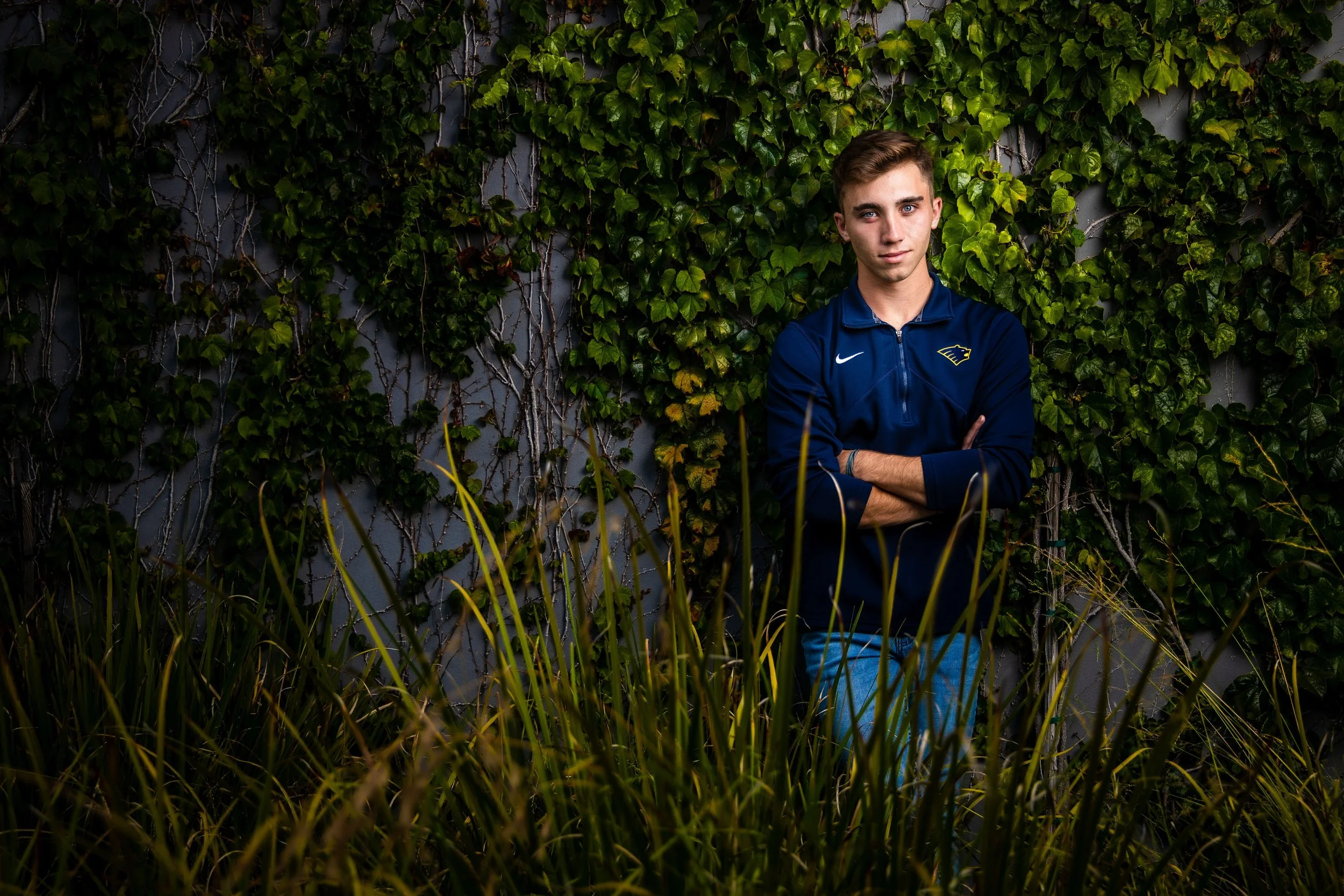 A young man stands with arms crossed in front of a gray wall covered with green ivy leaves, surrounded by tall grass in the foreground.