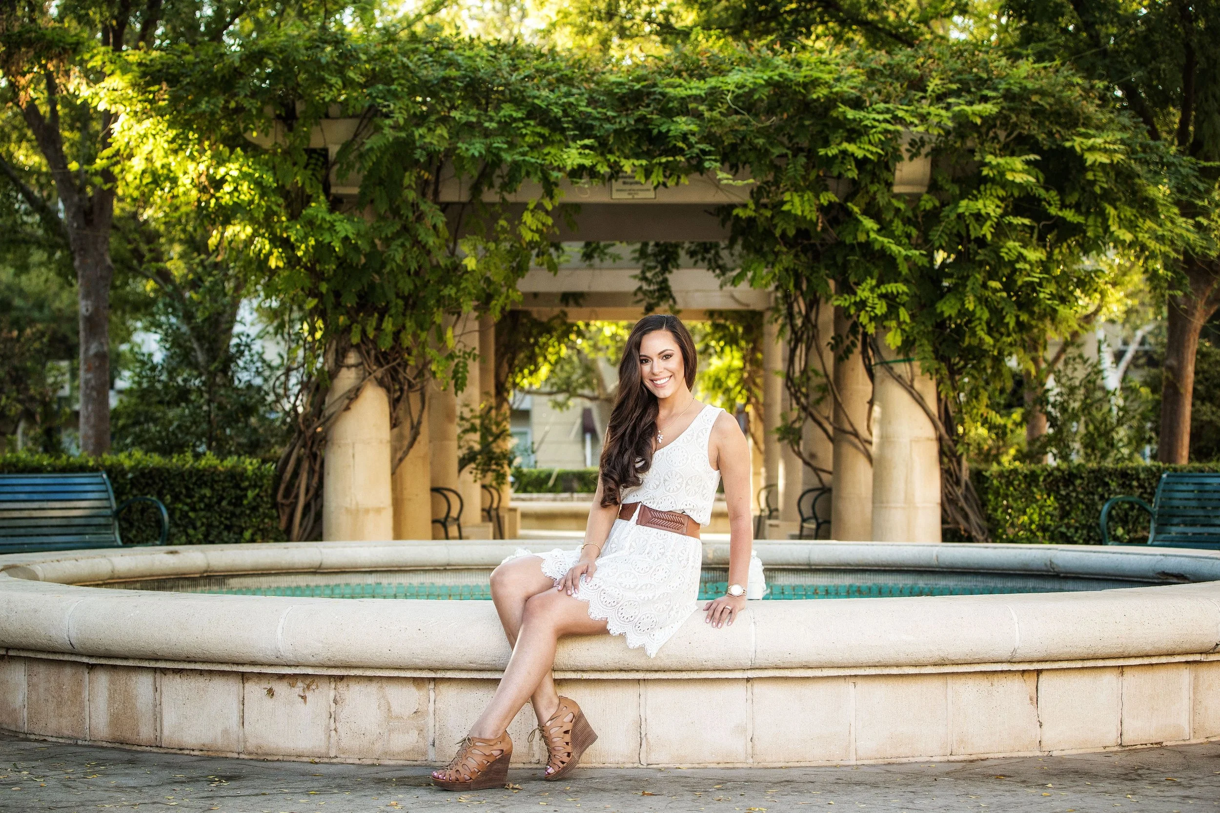 A smiling woman sitting on the edge of a fountain in a park with green trees and foliage in the background.