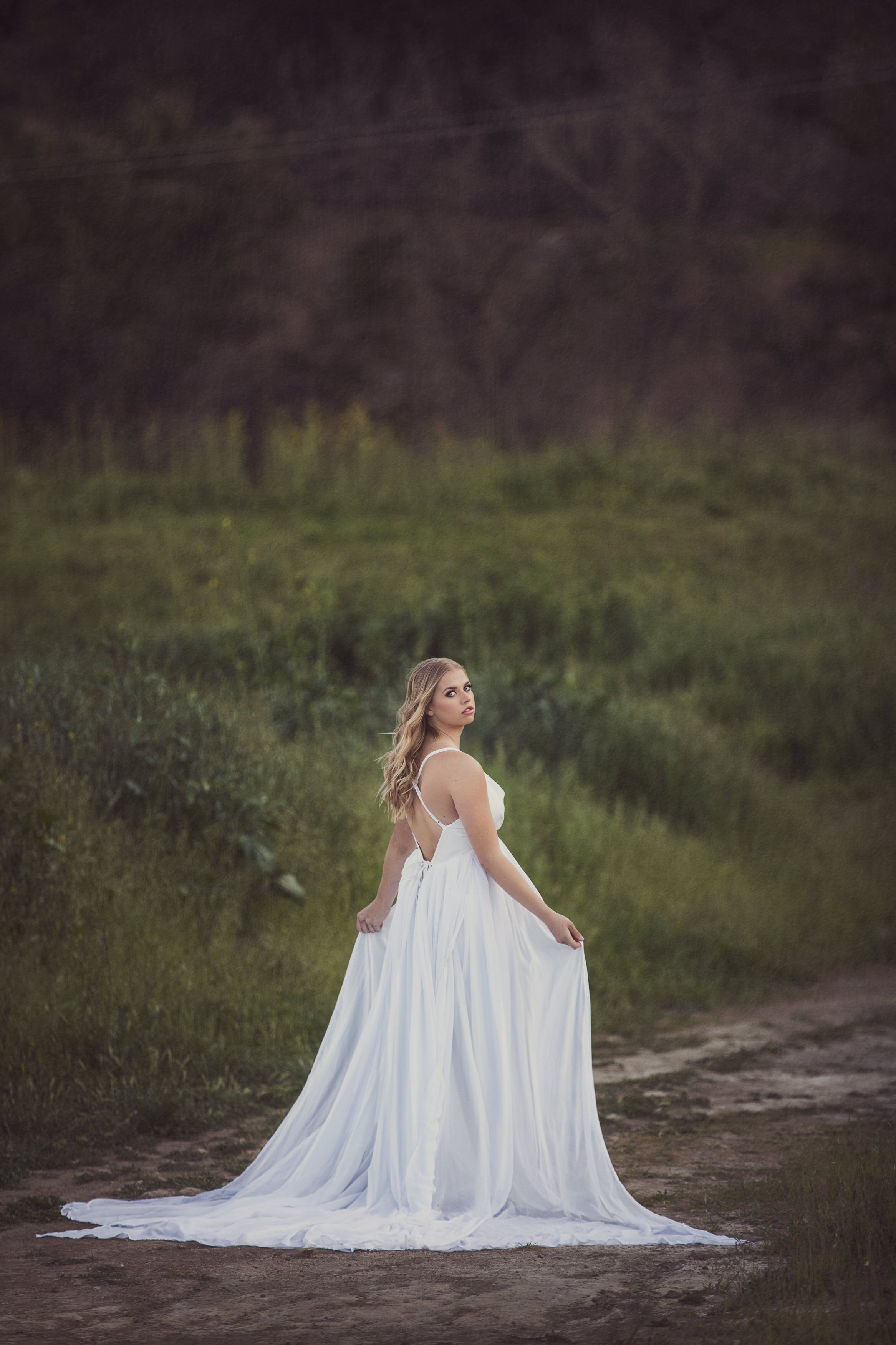 A woman in a white flowing dress standing on a dirt path in a rural outdoor setting with green grass and trees in the background.