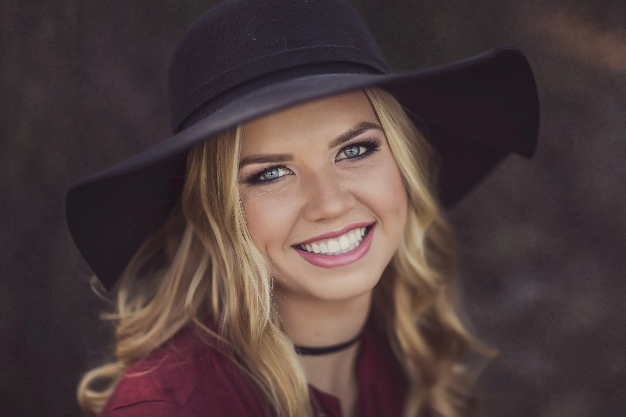 A woman with blonde hair, blue eyes, and a big smile wearing a large black floppy hat and a dark red top, outdoors against a blurred background.