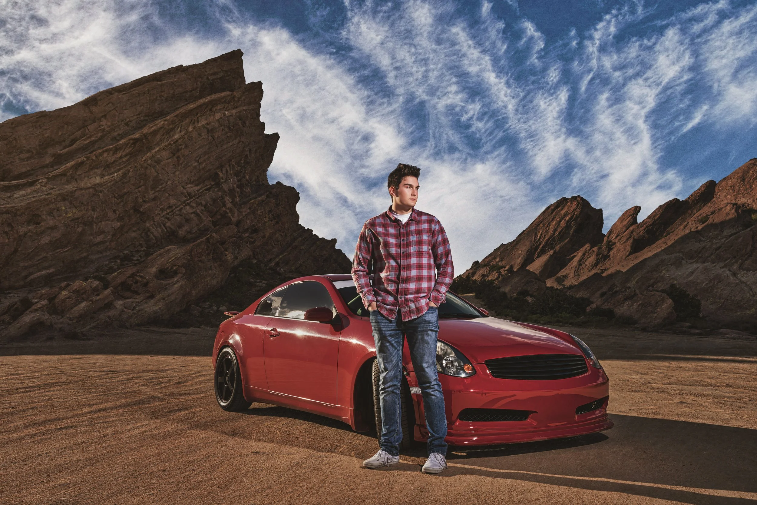 Young man standing in front of a red car on a dirt road with rocky mountains and a blue sky with wispy clouds in the background.