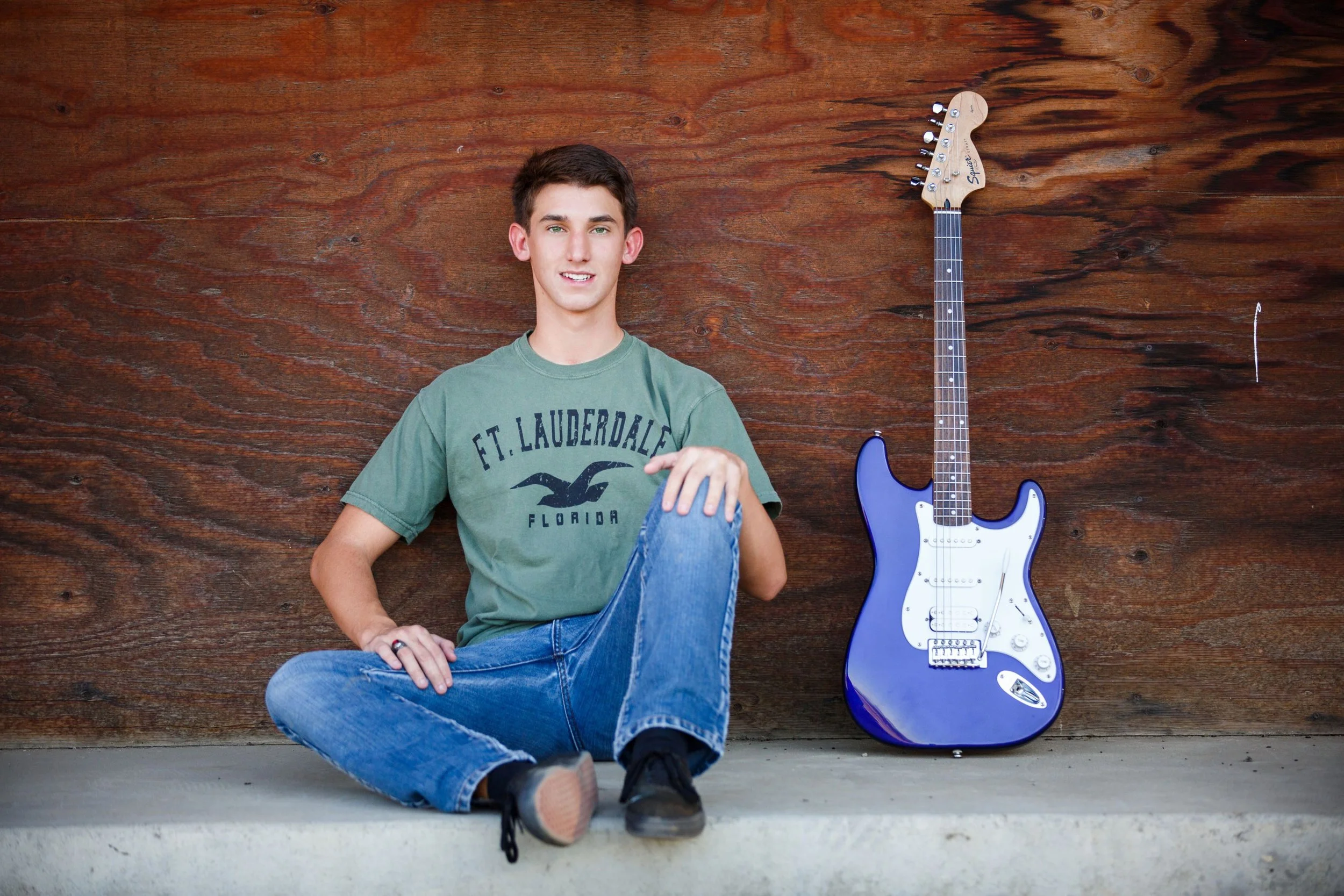 A young man sitting on the ground with a wooden wall behind him, next to an electric guitar leaning against the wall.