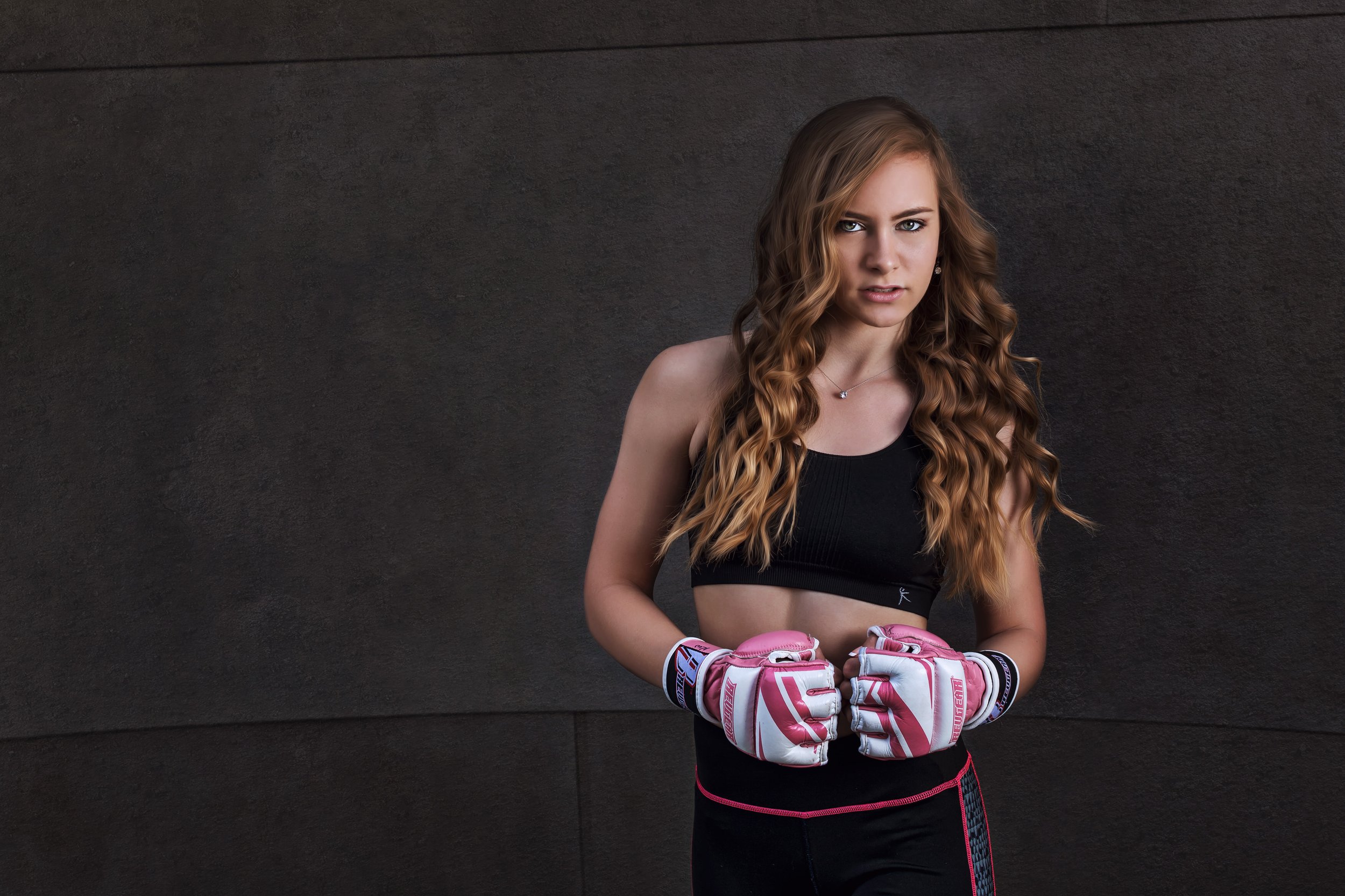 A young woman with long, wavy, reddish-brown hair wearing a black sports bra, black athletic shorts with pink accents, and pink and white gloves, standing against a textured dark background, looking directly at the camera with a determined expression